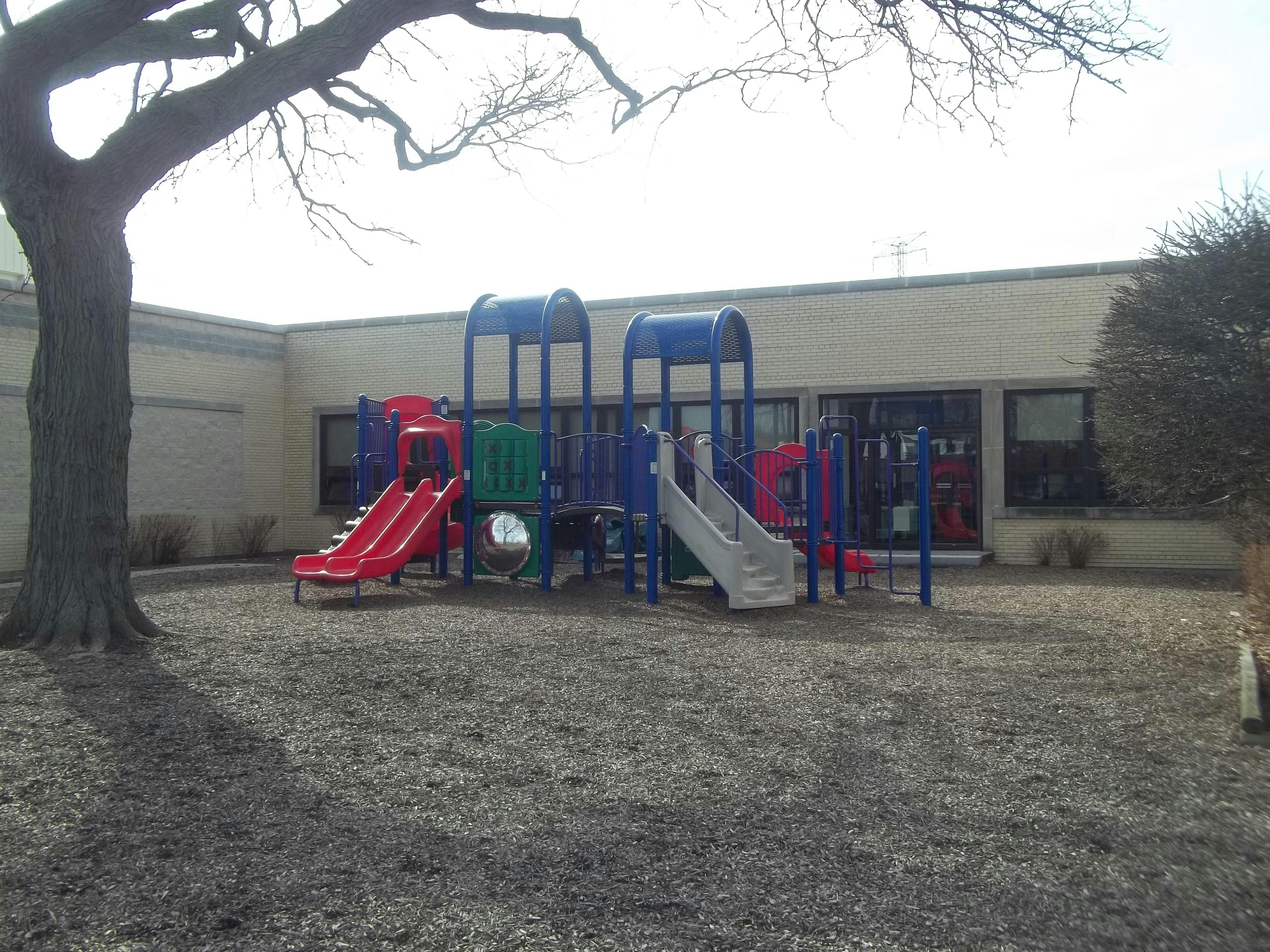 Playground with slides and climbing structures on wood chips next to a building