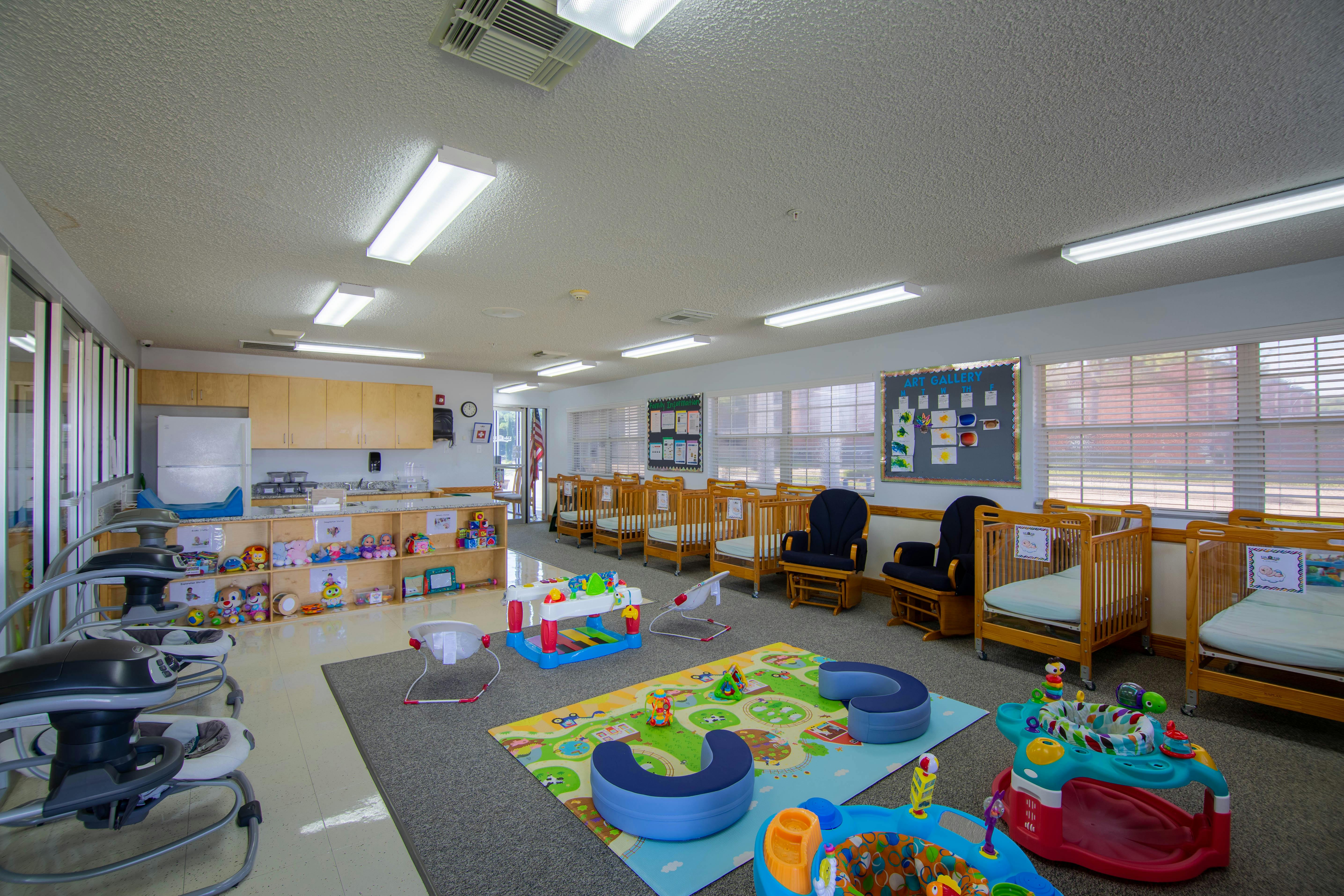 A brightly lit nursery room with cribs, playmats, infant swings, and a changing station