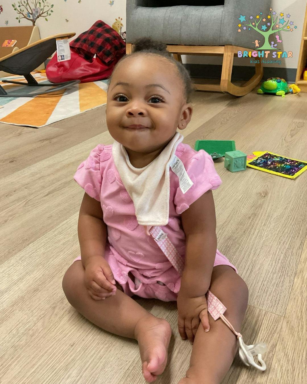 Child in pink outfit and bib sitting on a wooden floor