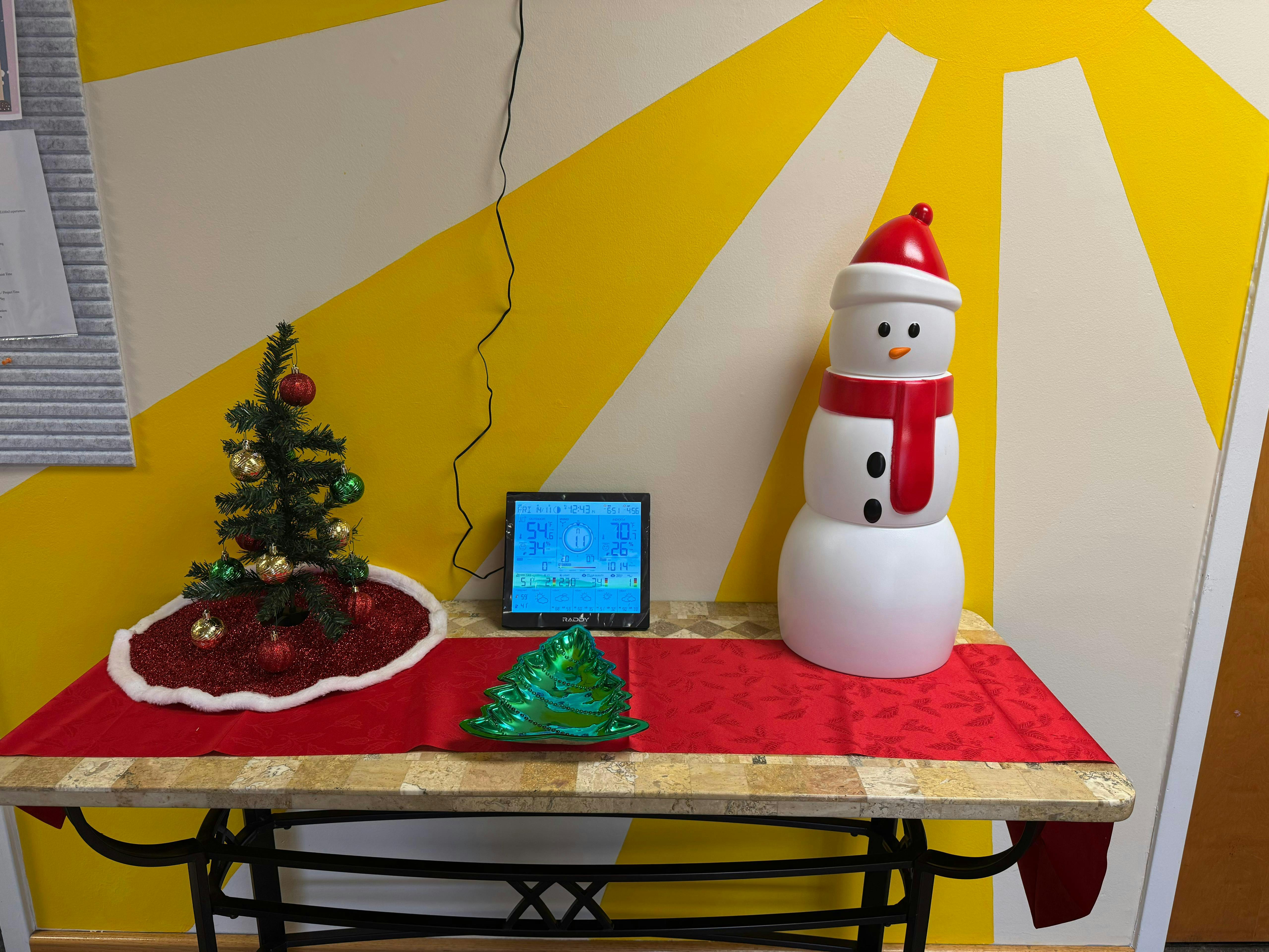 Small decorated Christmas tree, a digital weather display, and a snowman figure on a table