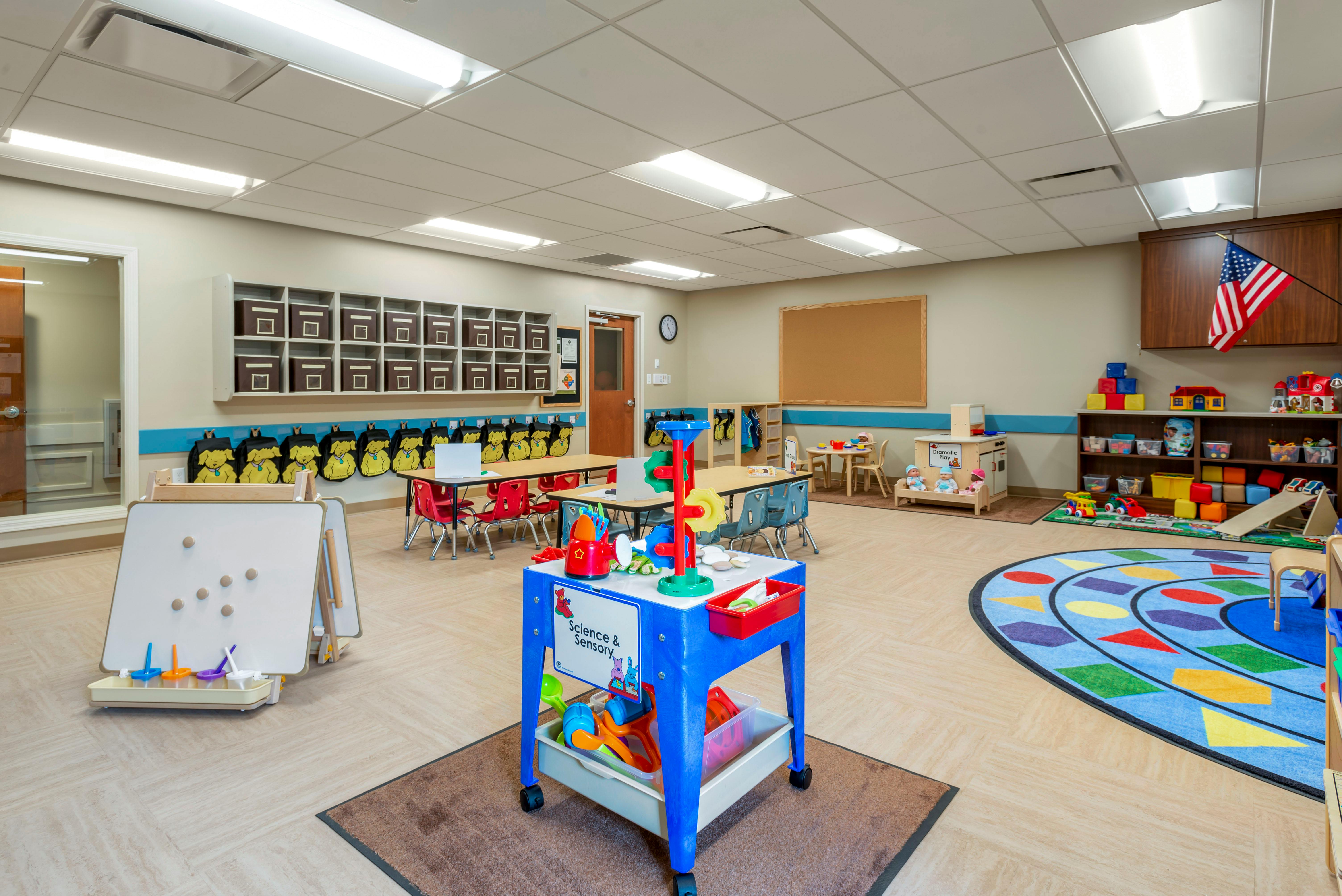 Empty classroom with activity tables, cubbies, and a colorful rug