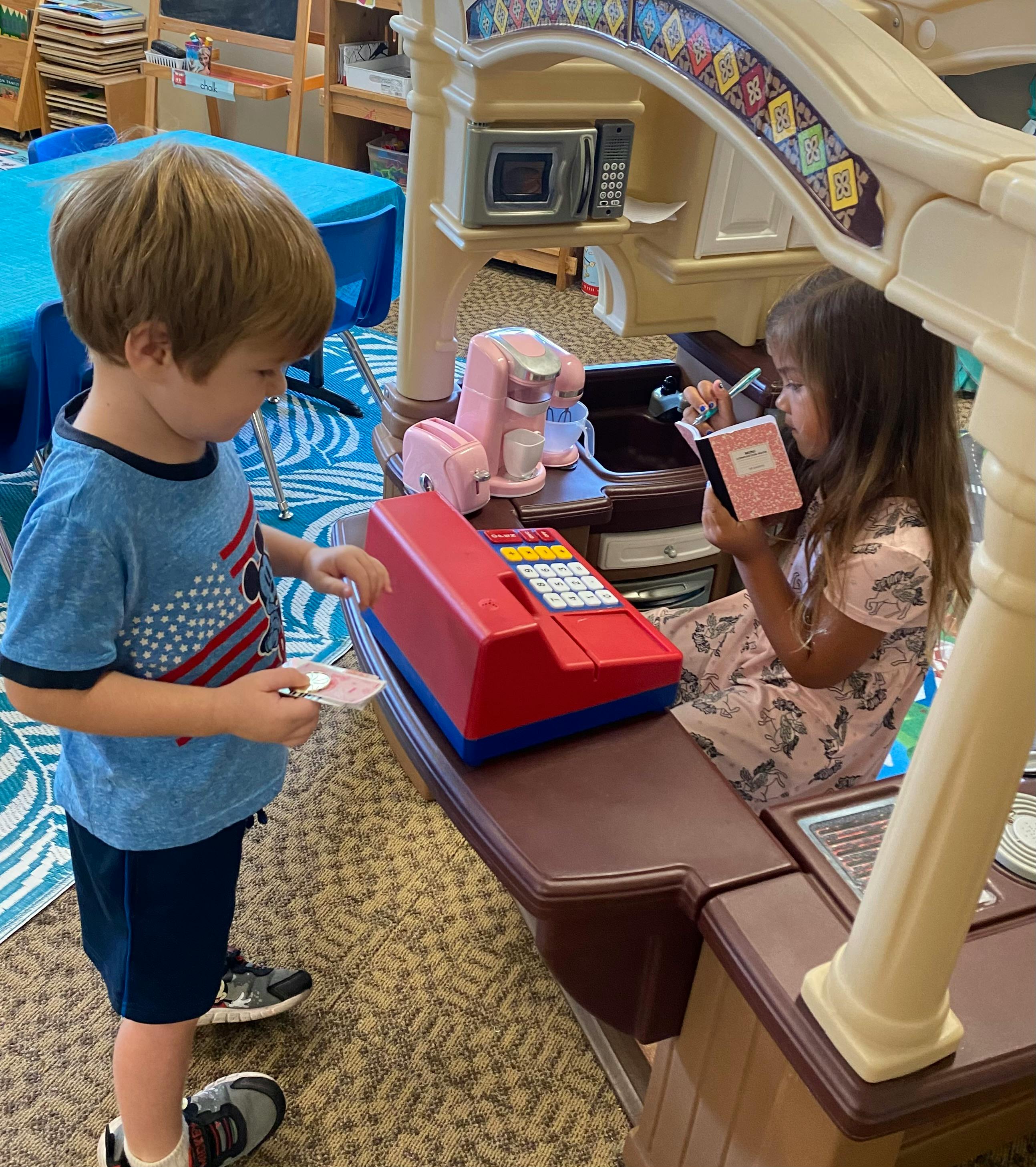 Child holding a coin at a toy register with another child reading a book