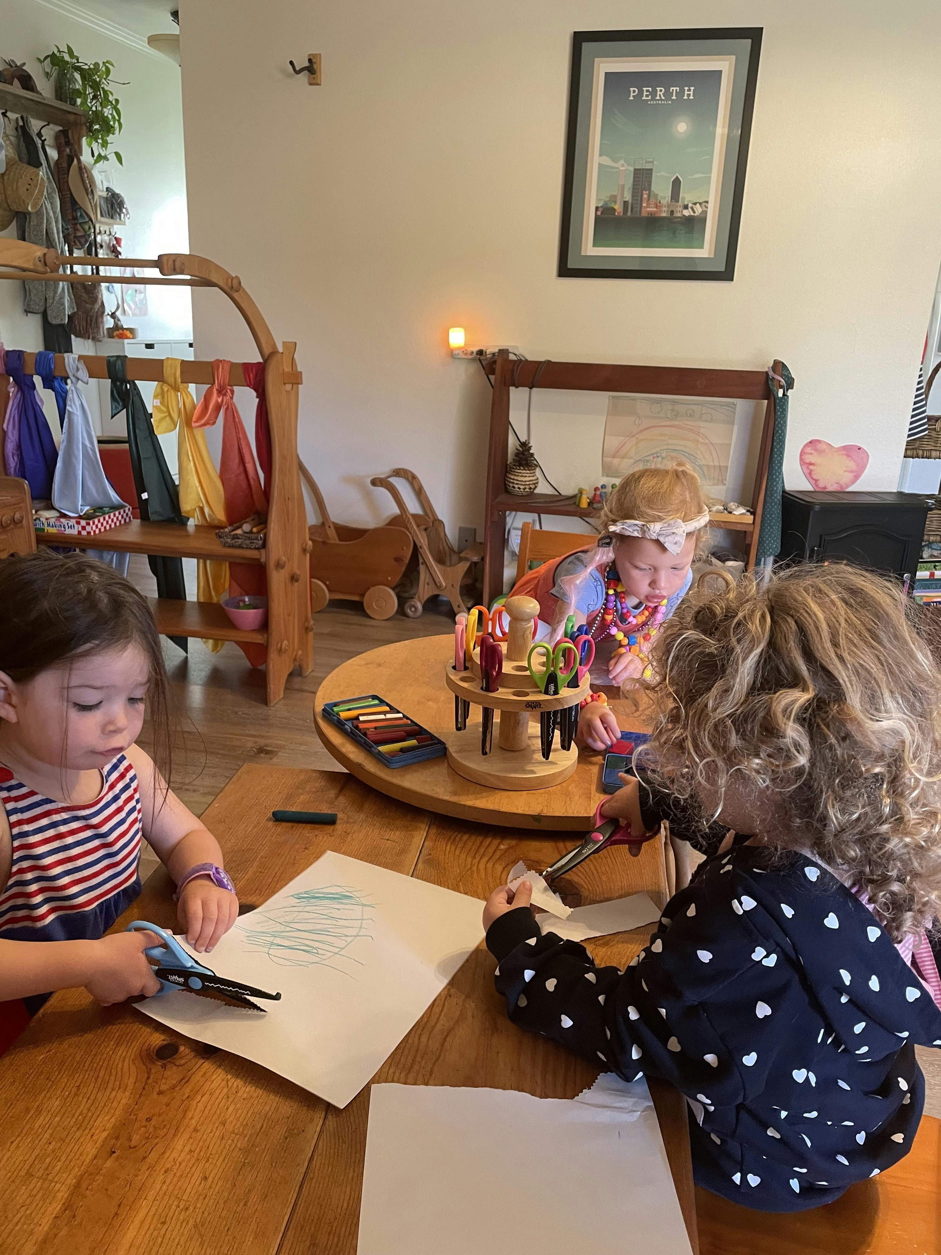 Three young children cutting paper and drawing with crayons at a wooden table