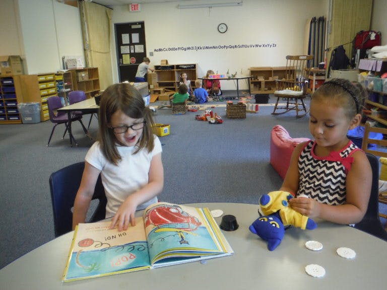 Two children at a table in a classroom, one reading a book, one playing with a toy