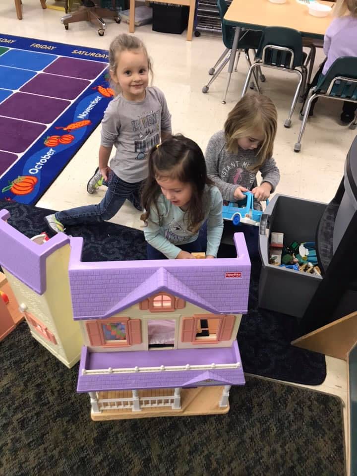 Three children playing with a dollhouse and toys in a classroom