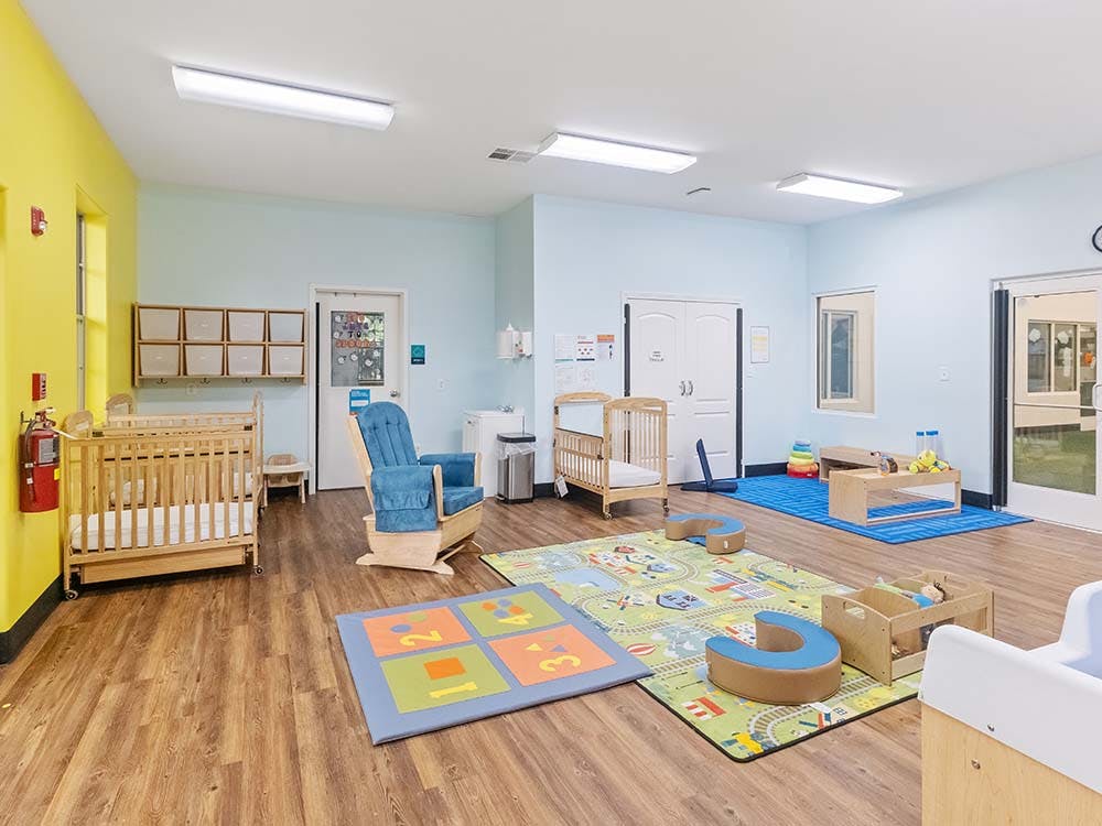 A colorful childcare room with cribs, playmats, and a rocking chair
