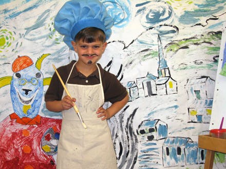 Child in chef hat and apron with painted mustache holding a paintbrush