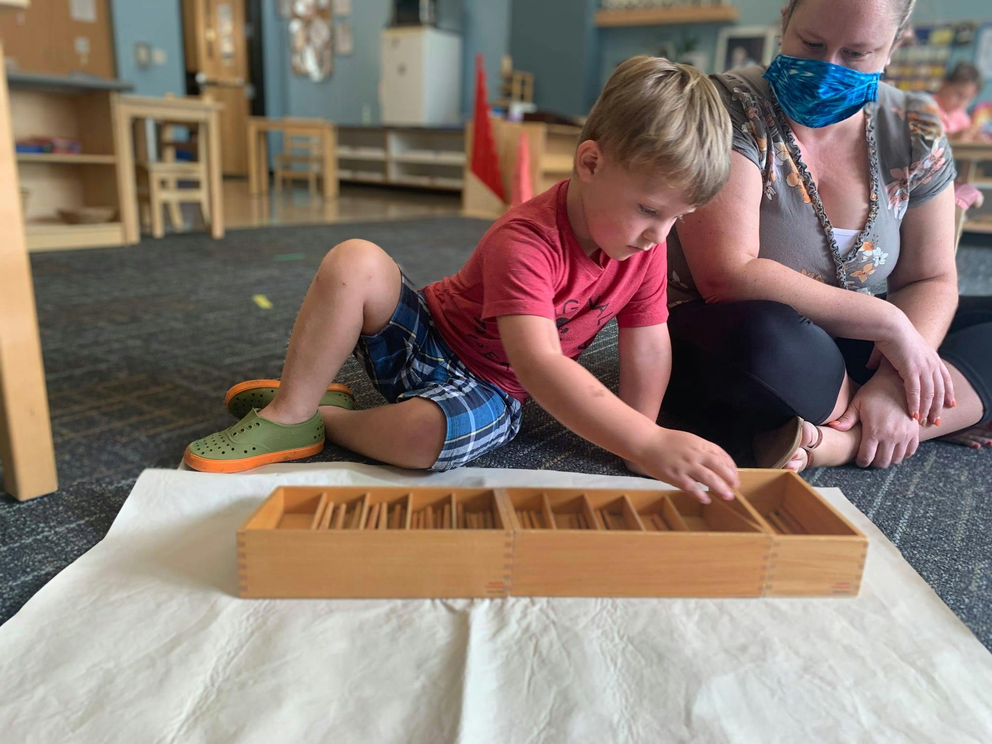 Child and person in mask on floor with wooden sorting tray