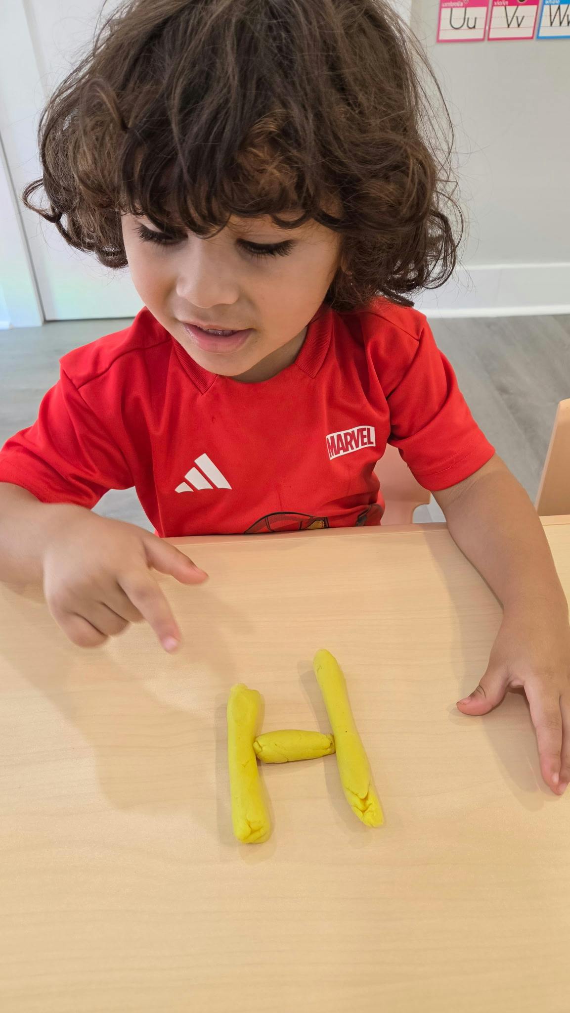 Child pointing at the letter H made from yellow modeling clay