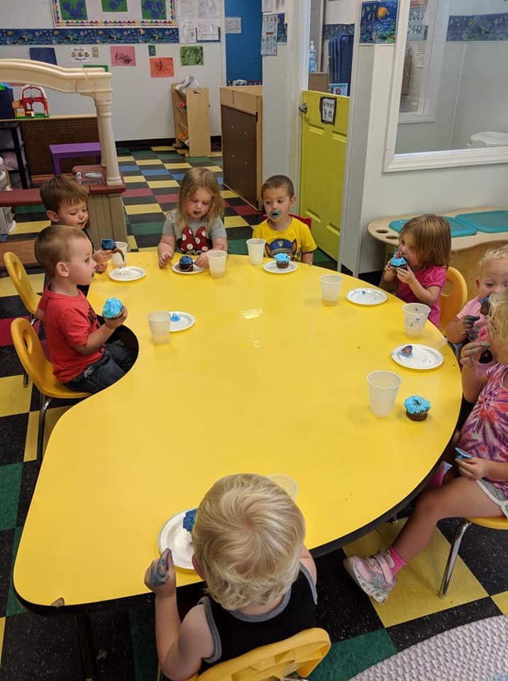 Children sitting around a yellow table eating cupcakes in a classroom