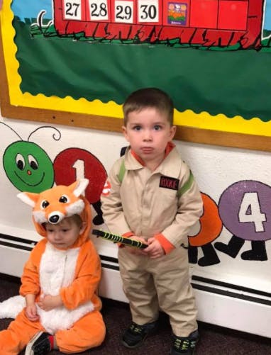 Child in fox costume sitting next to child in tan jumpsuit holding a toy