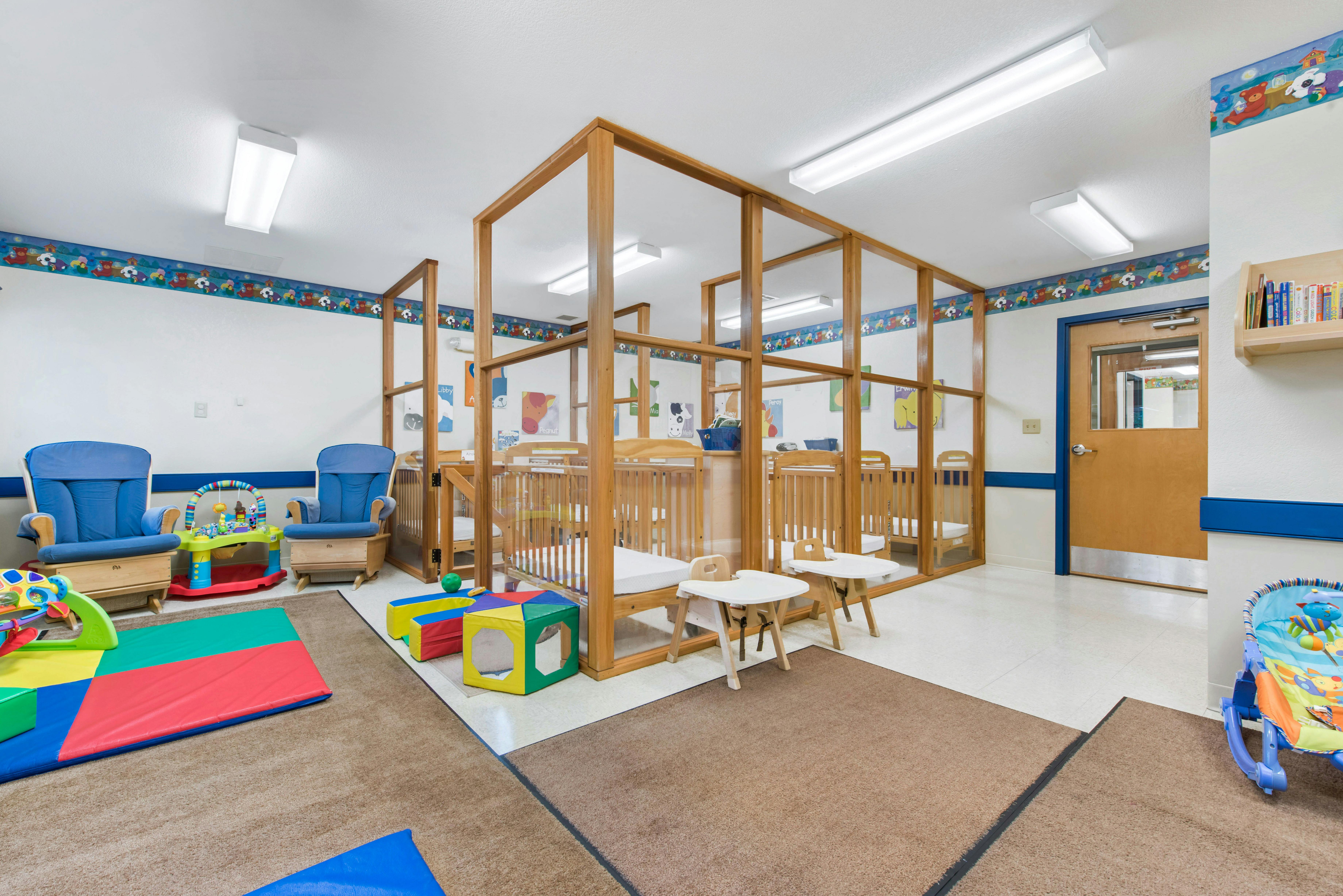 A room with multiple wooden cribs separated by dividers, blue rocking chairs, and toys