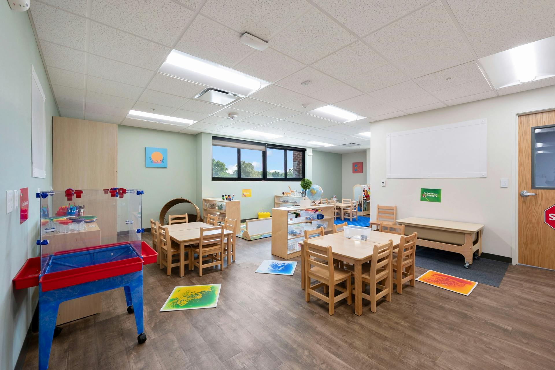 Classroom with small wooden tables and chairs, activity centers, and large windows