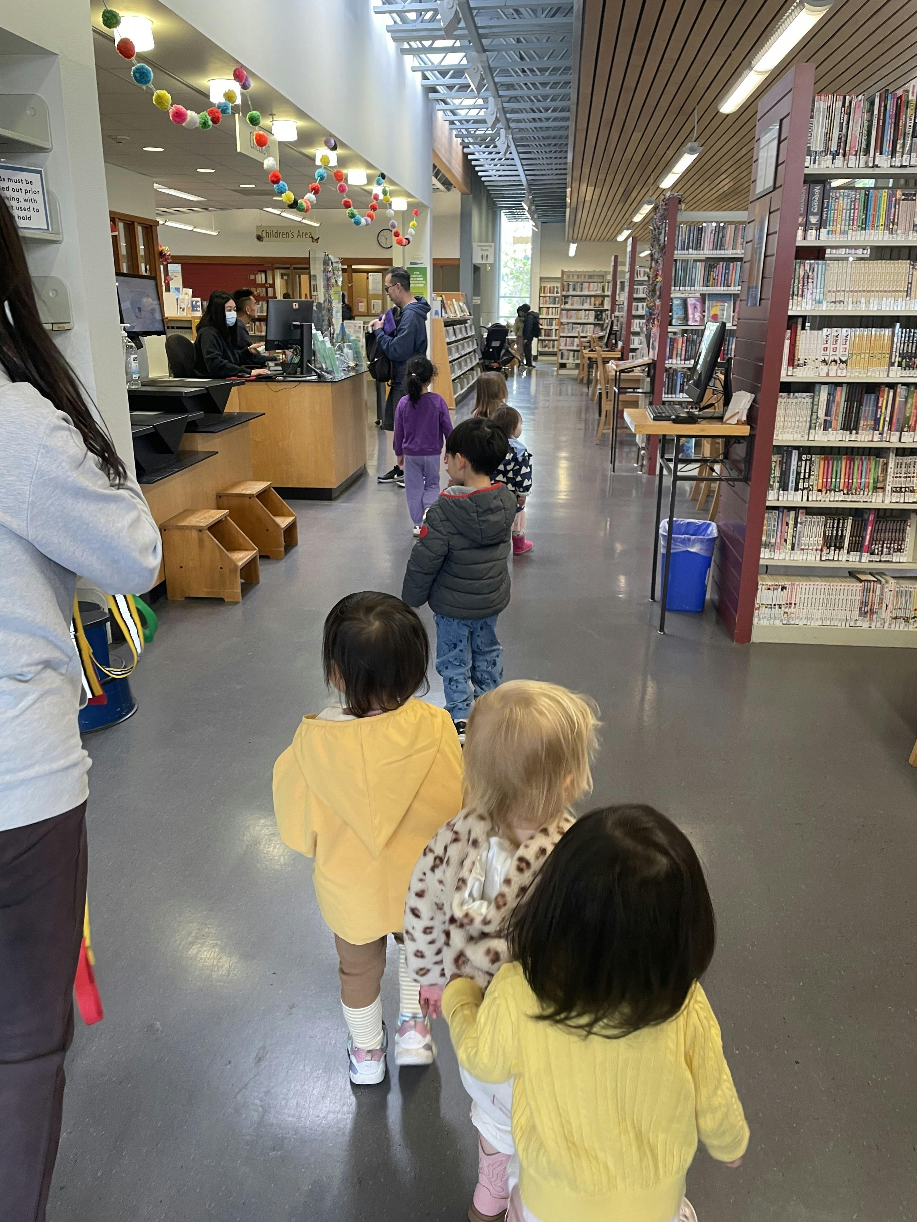Children walking in a line through a library with shelves and service desk