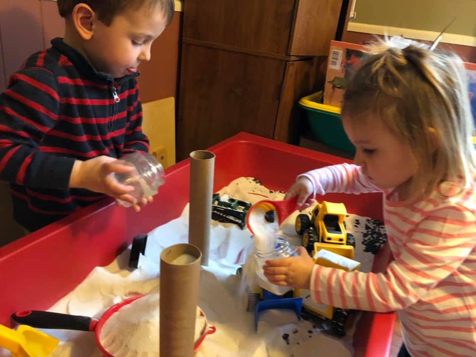 Two children playing in a red sensory bin with white rice and toys