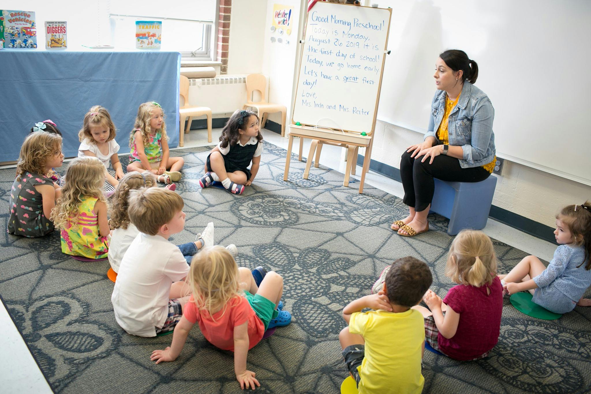 Person sitting on a stool facing a circle of children on the floor in a classroom