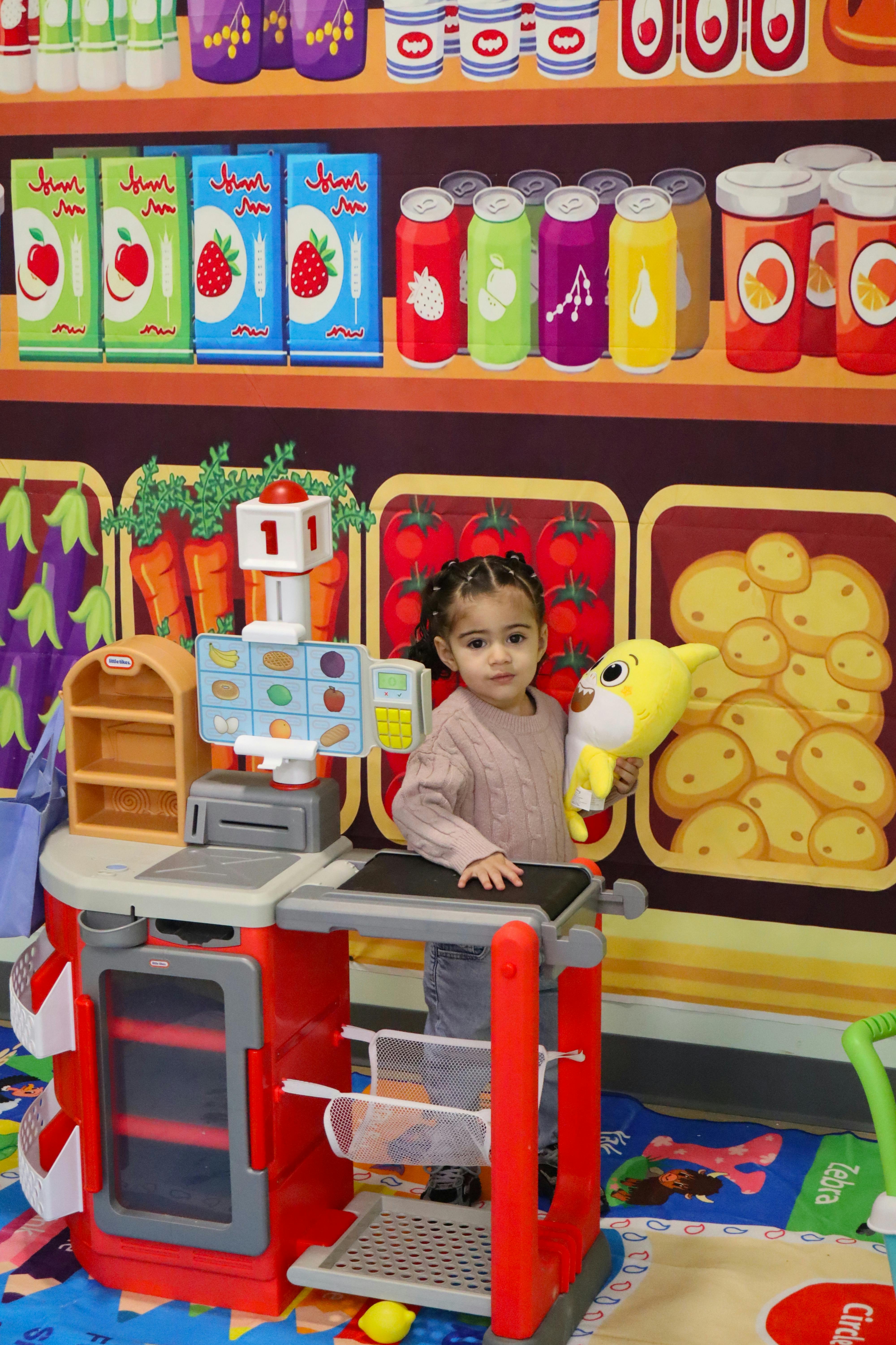 Child standing at a toy checkout counter holding a yellow stuffed animal