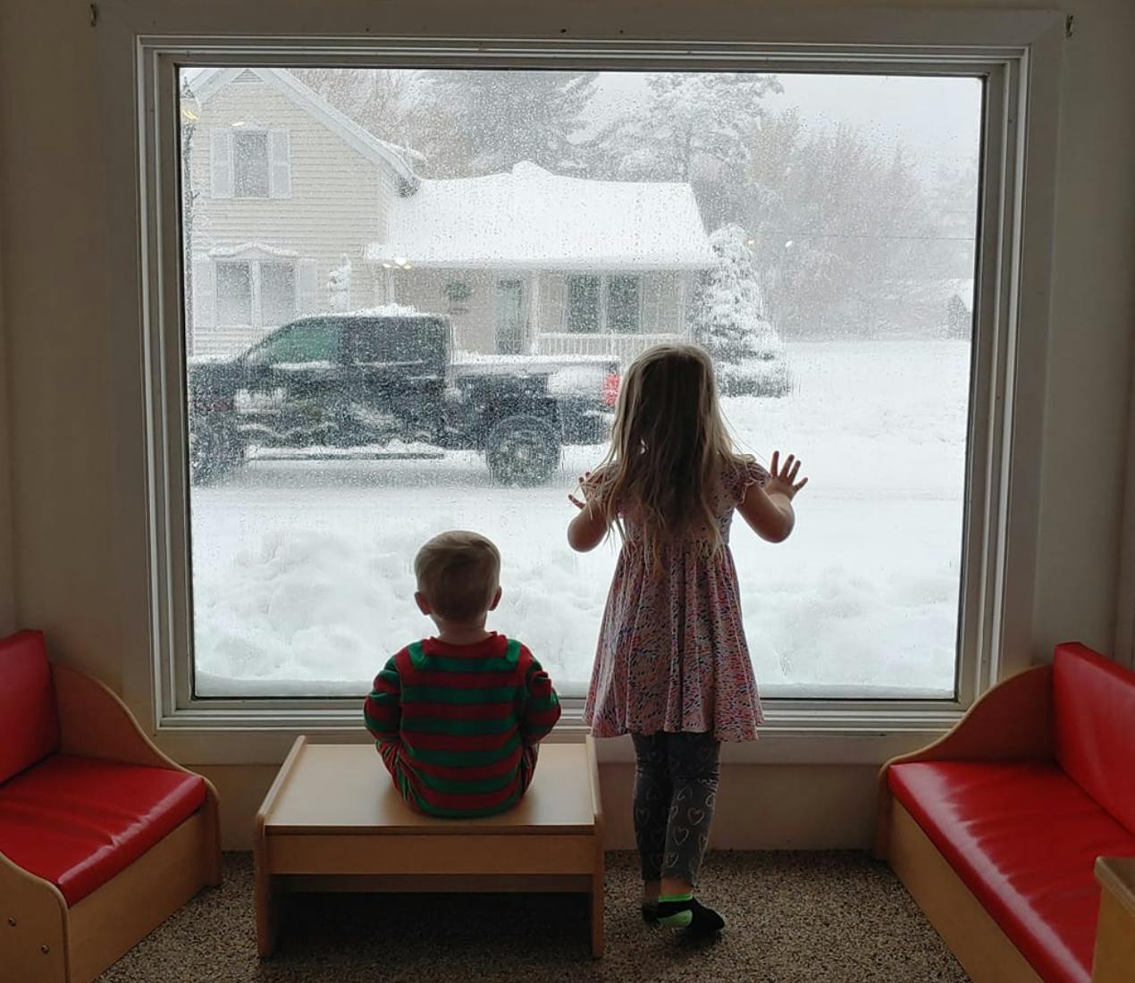 Two children looking out a window at a snowy street with a truck and house