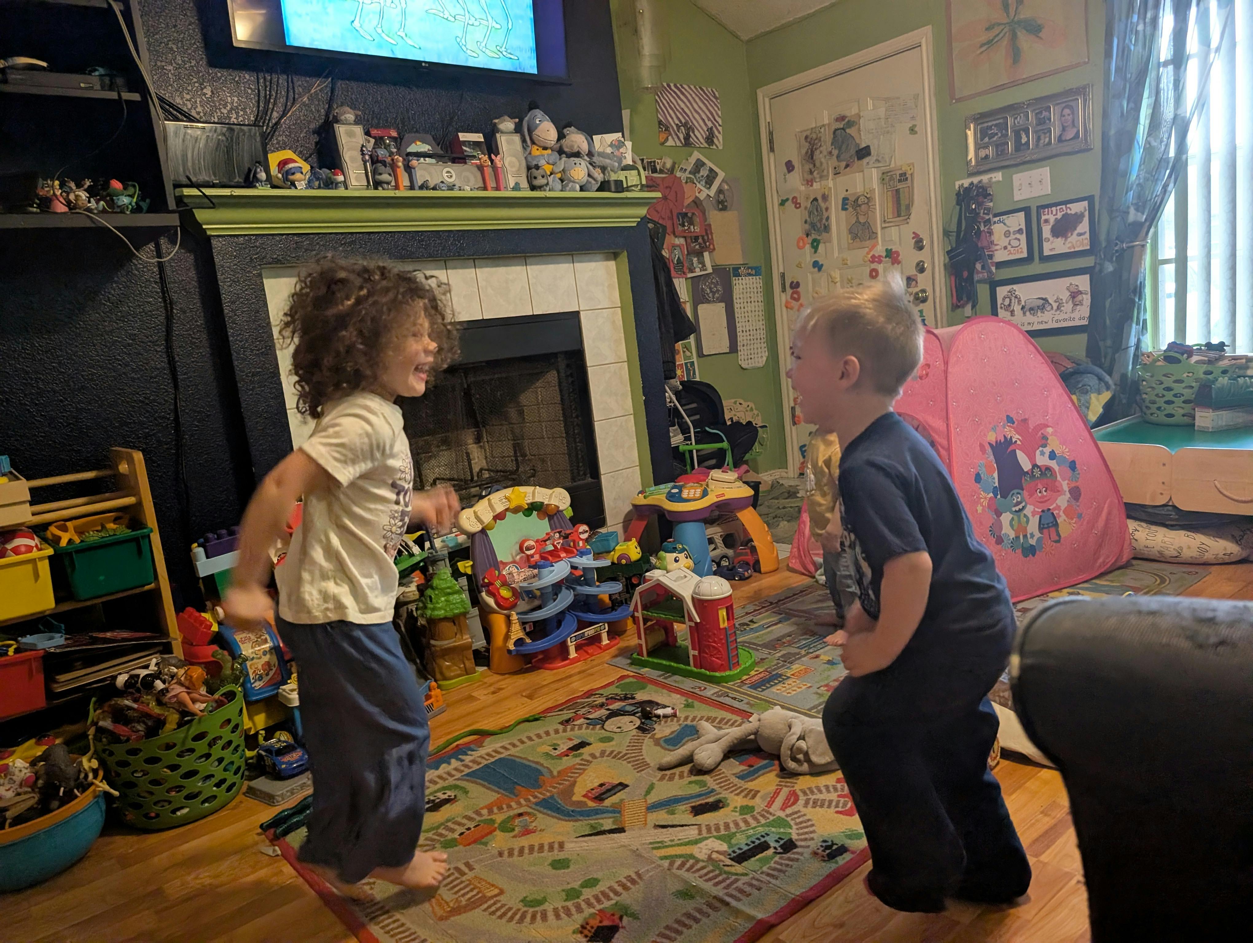 Two children playing in a living room filled with toys, smiling at each other