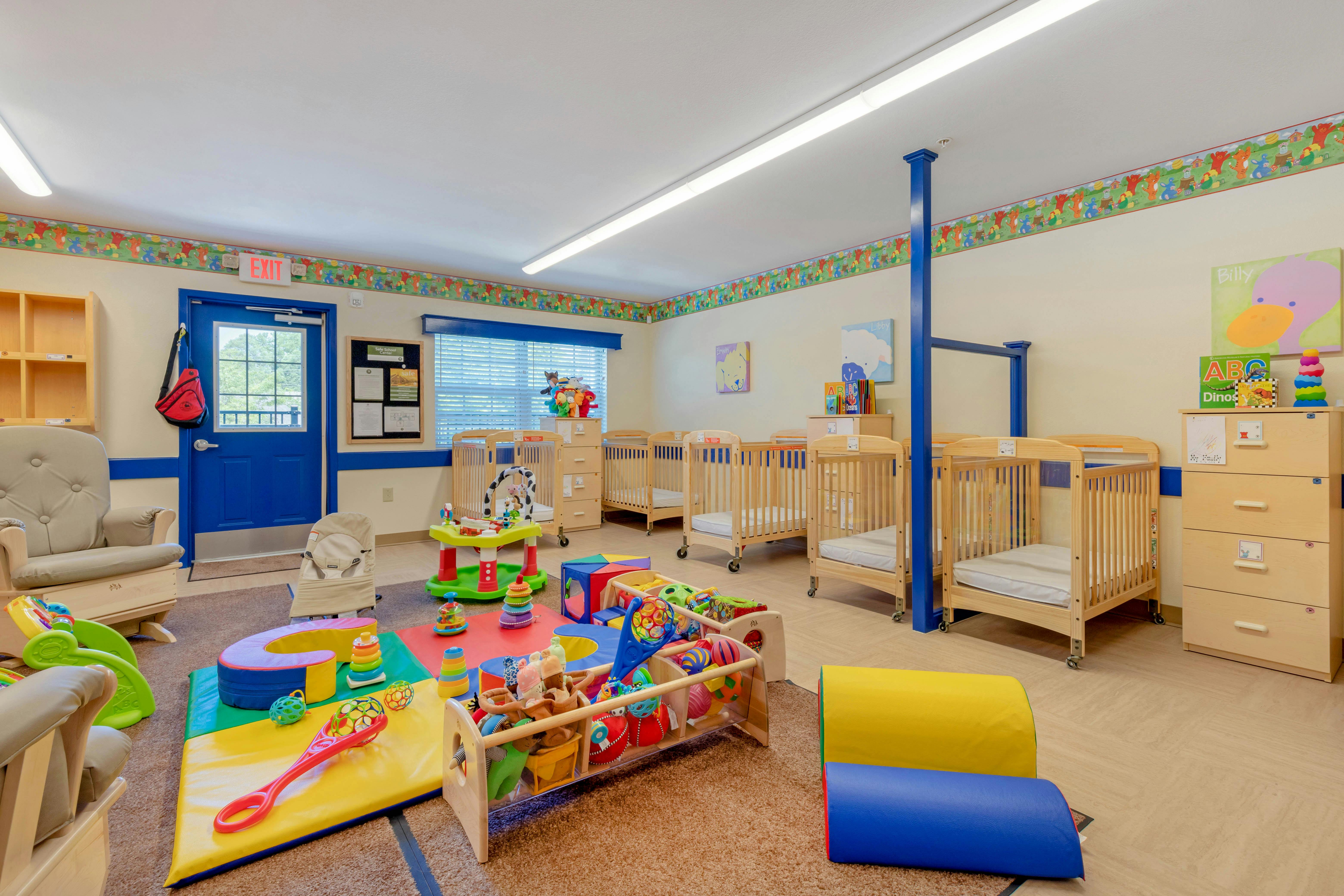 A classroom with cribs, play mats, and toys, with an exit sign over a blue door
