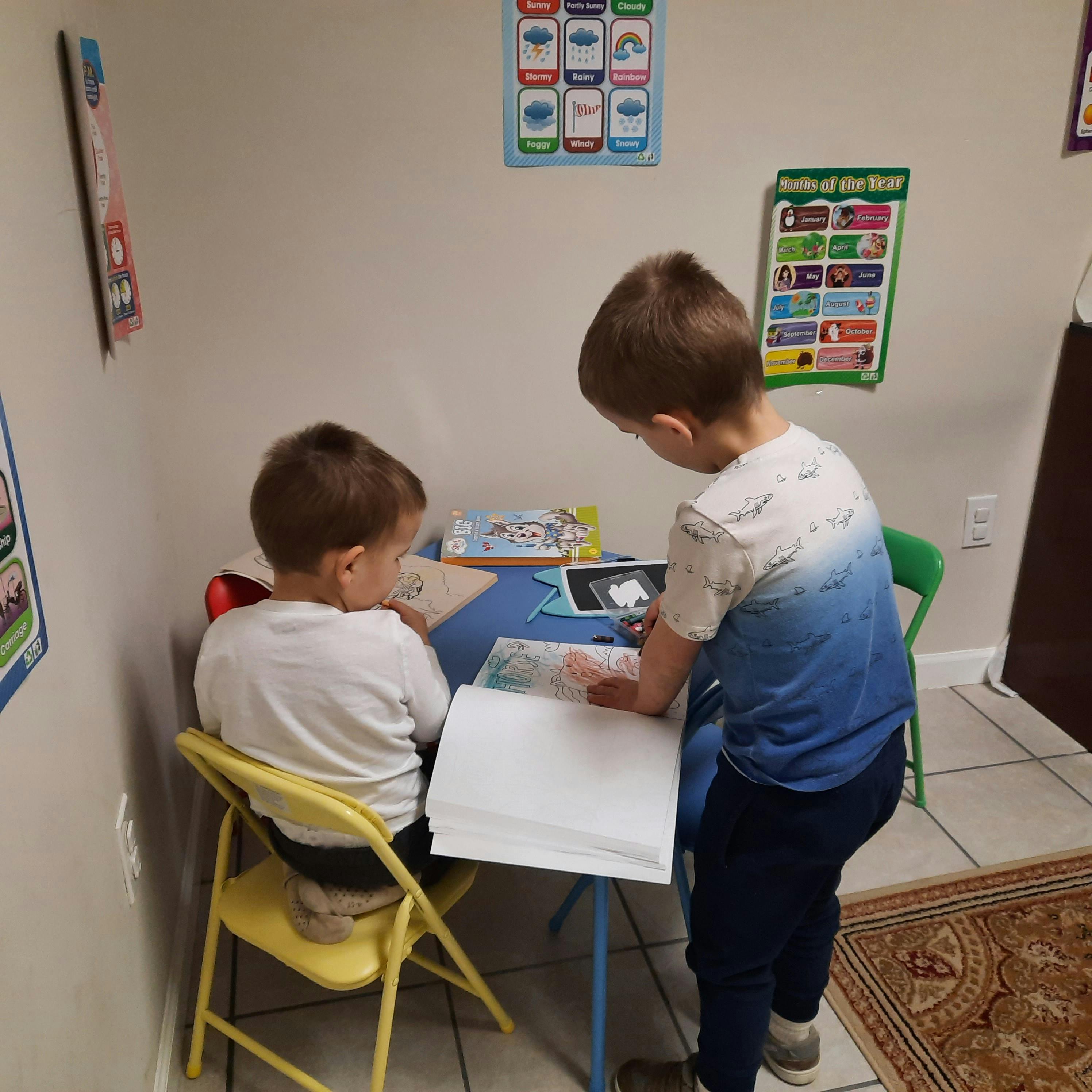 Two children at a table, one seated and one standing, looking at papers