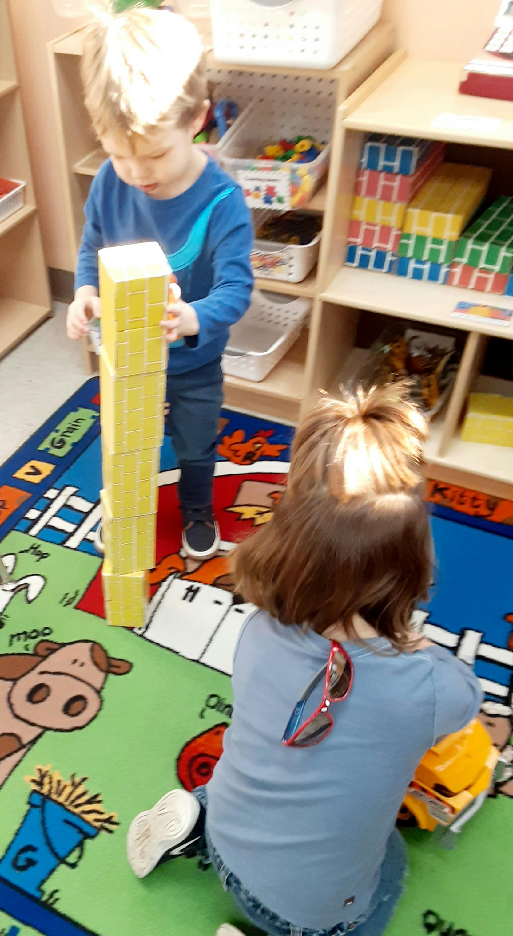 Child building a tall tower with yellow blocks, another child playing nearby