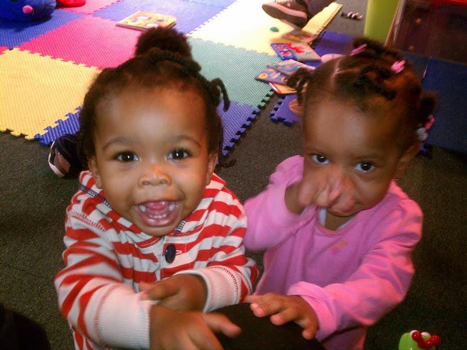 Two children on a colorful mat, one smiling and one with a finger near their nose