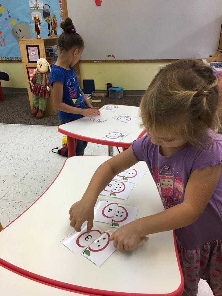 Two children working with apple-themed number puzzles at tables in a classroom