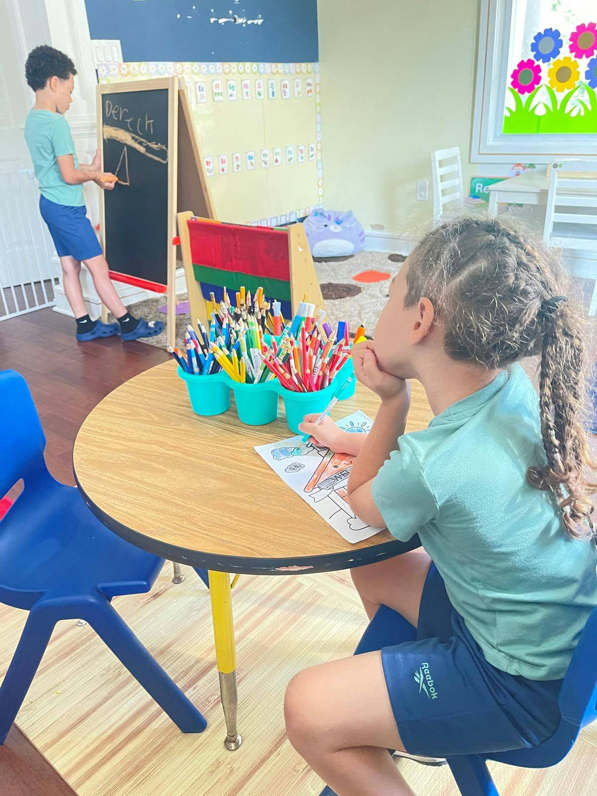 Child coloring at a table while another child writes on a blackboard