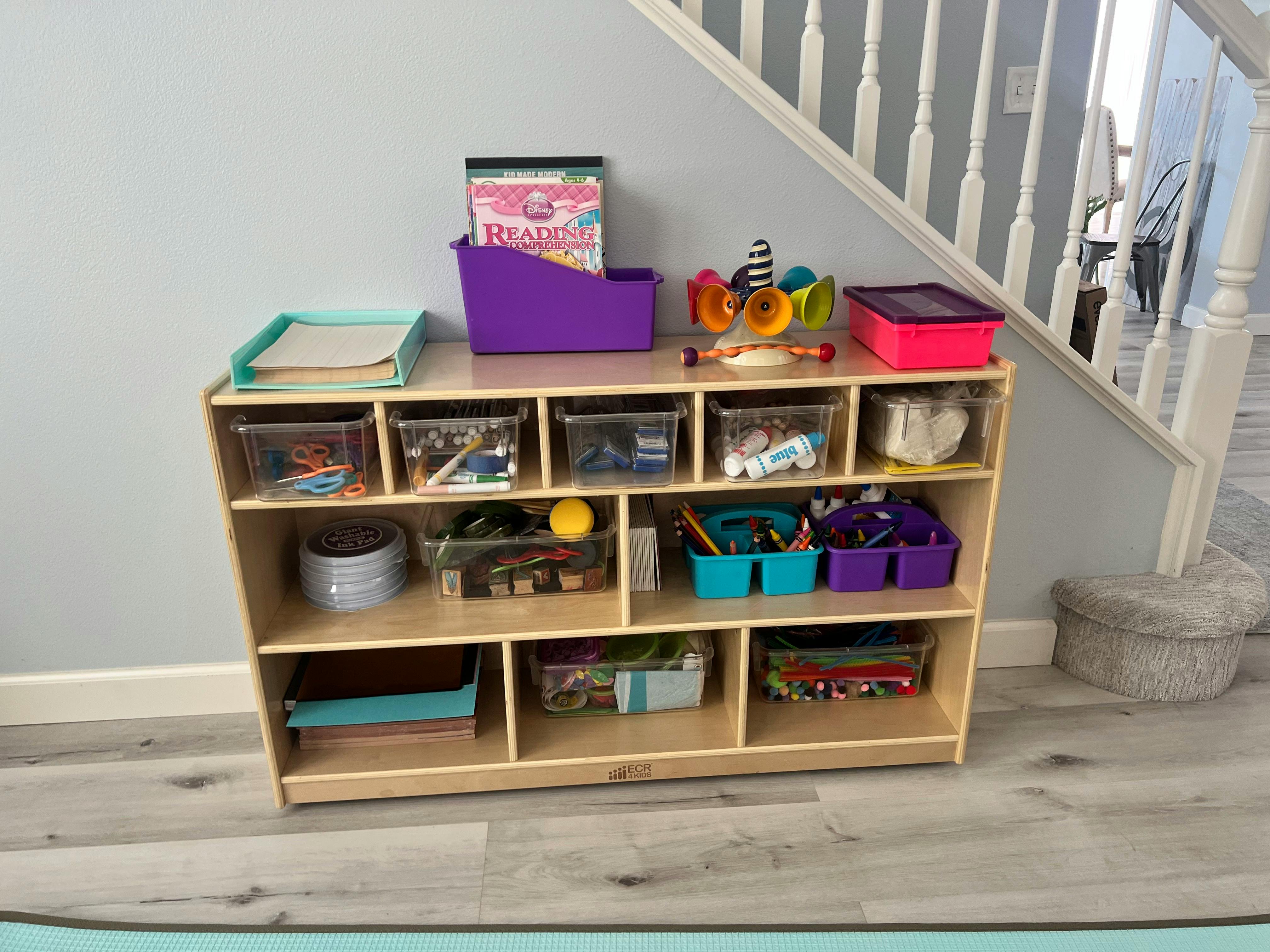 Wooden storage shelf with various bins and items under a staircase