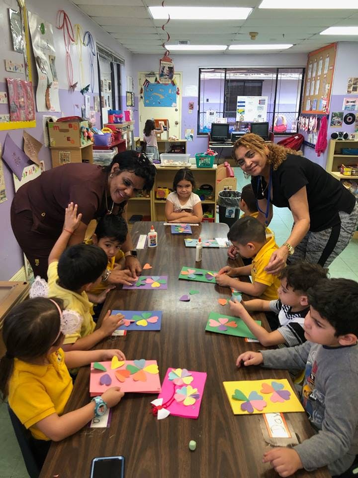 Two adults helping children with crafts at a table in a classroom