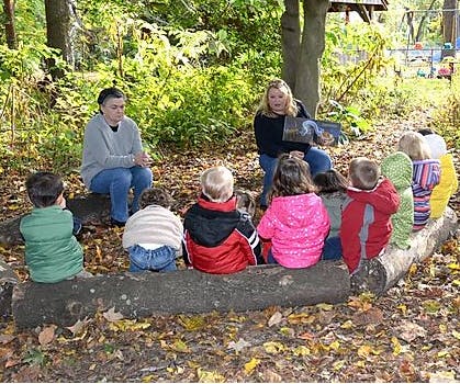 Two adults reading to a group of children sitting on a log outdoors