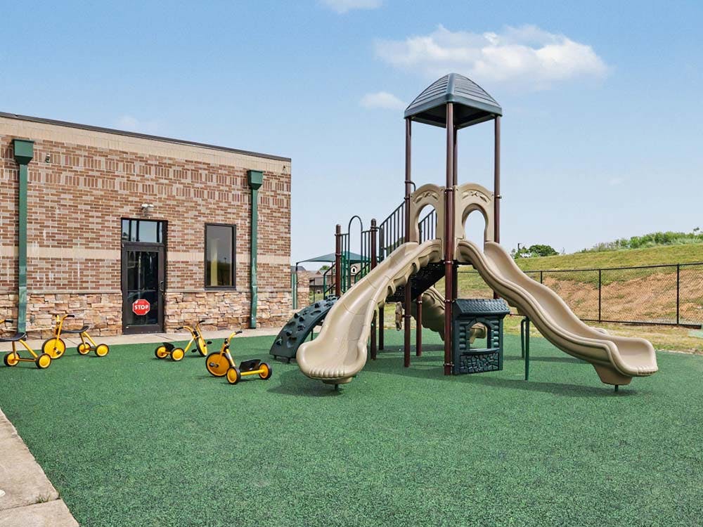Playground with slides and tricycles on green turf next to a brick building