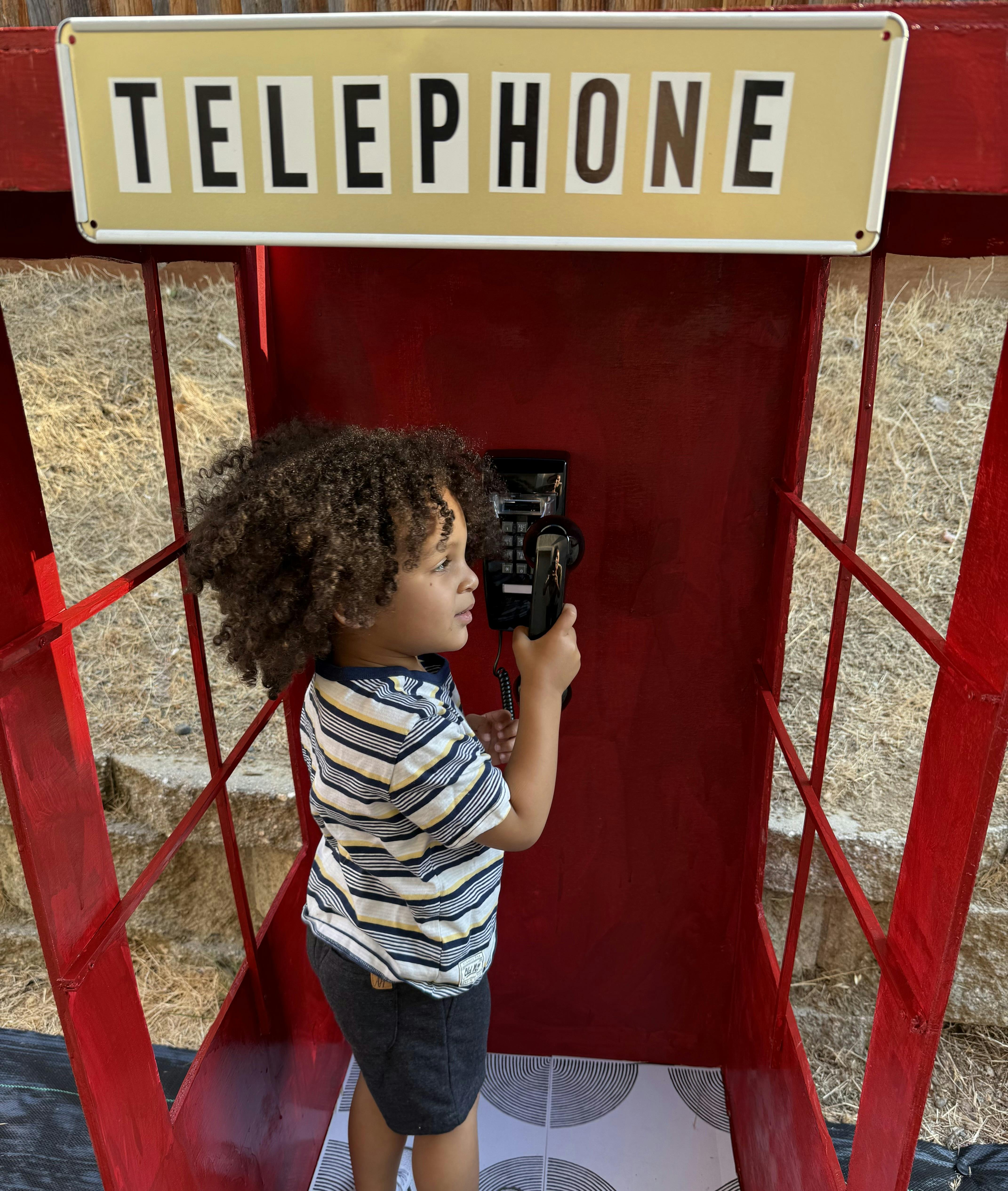Child holding a telephone receiver in a red telephone booth with a sign that reads "TELEPHONE"