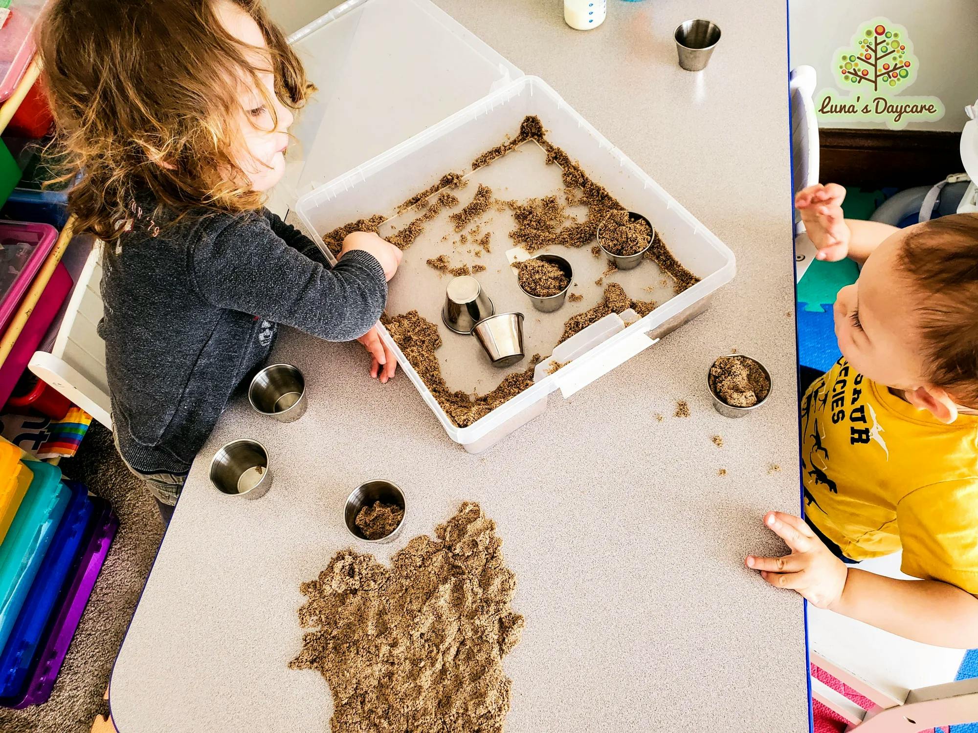 Two children playing with kinetic sand and small metal cups
