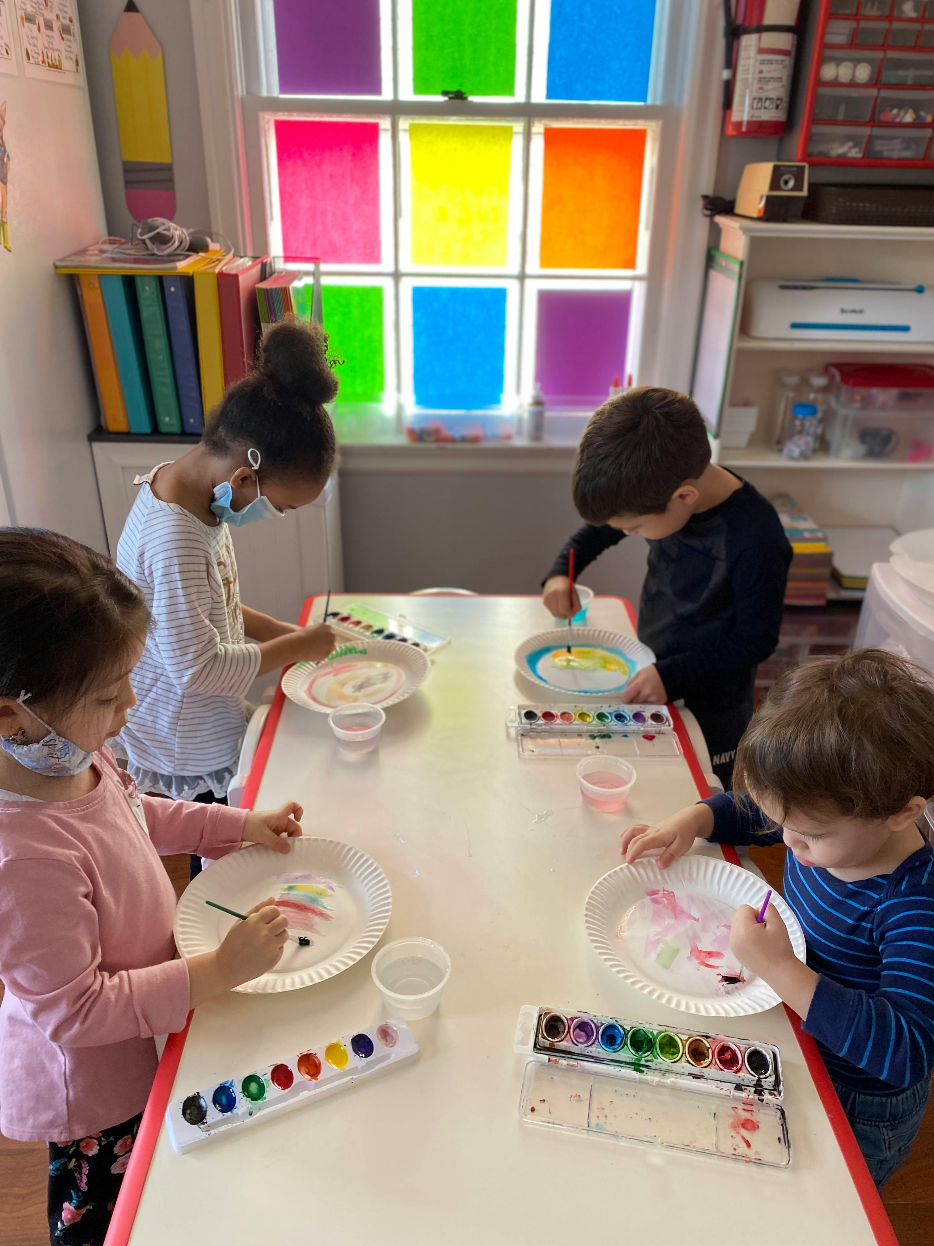Four children painting paper plates at a table in a classroom