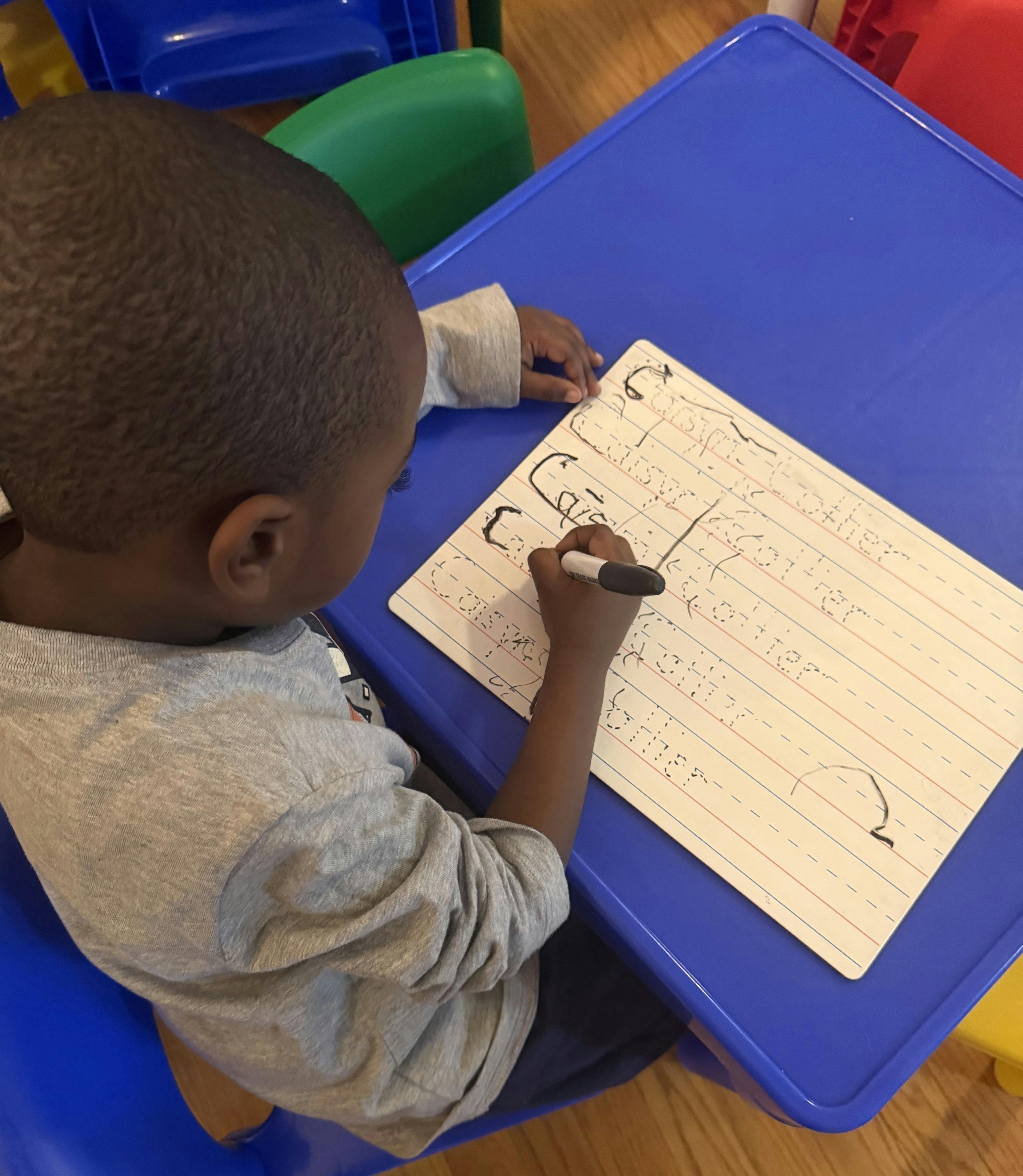 Child sitting at a blue table, writing with a marker on a white board