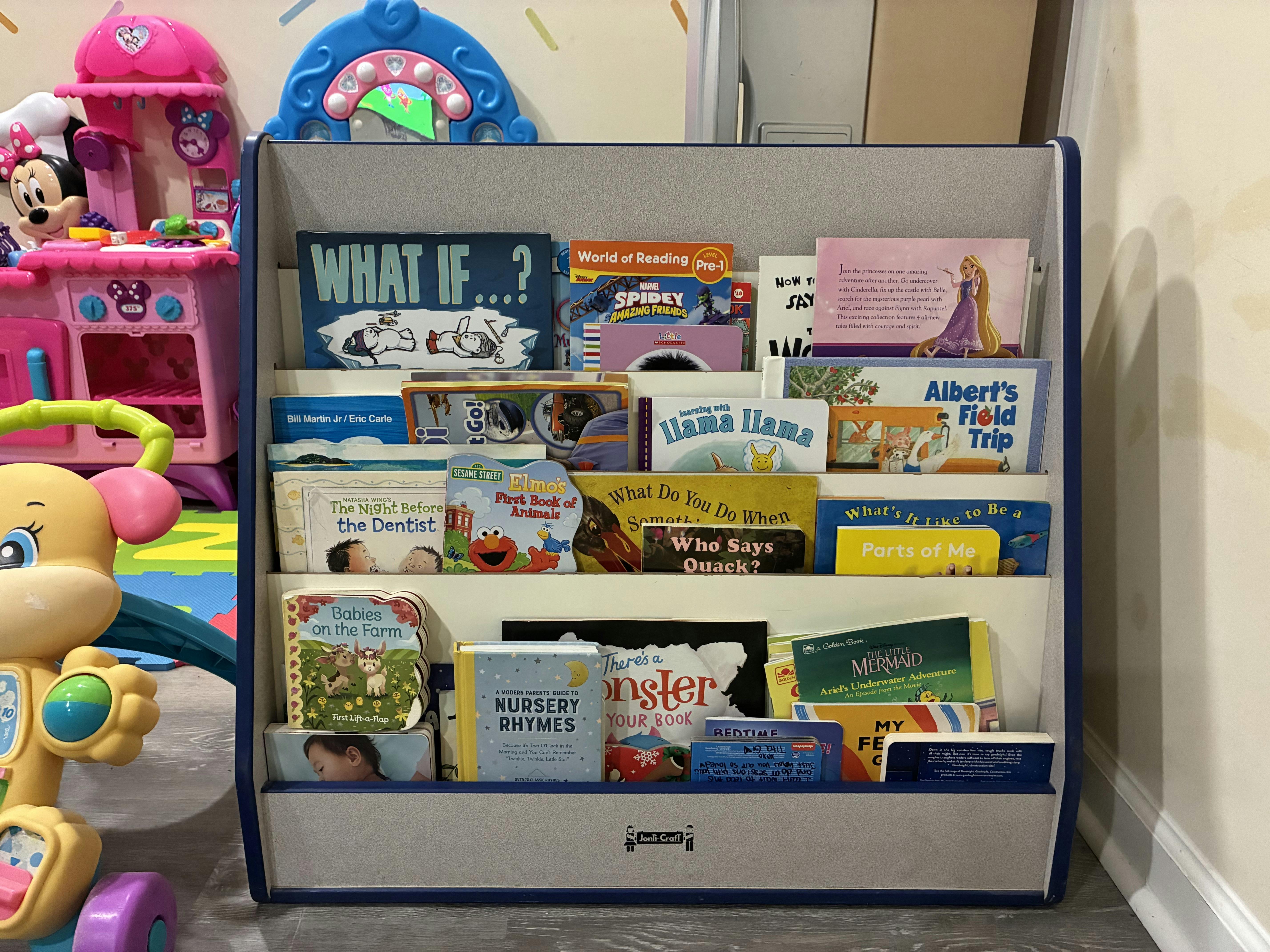 Children's books displayed in a grey bookstand next to a pink toy kitchen