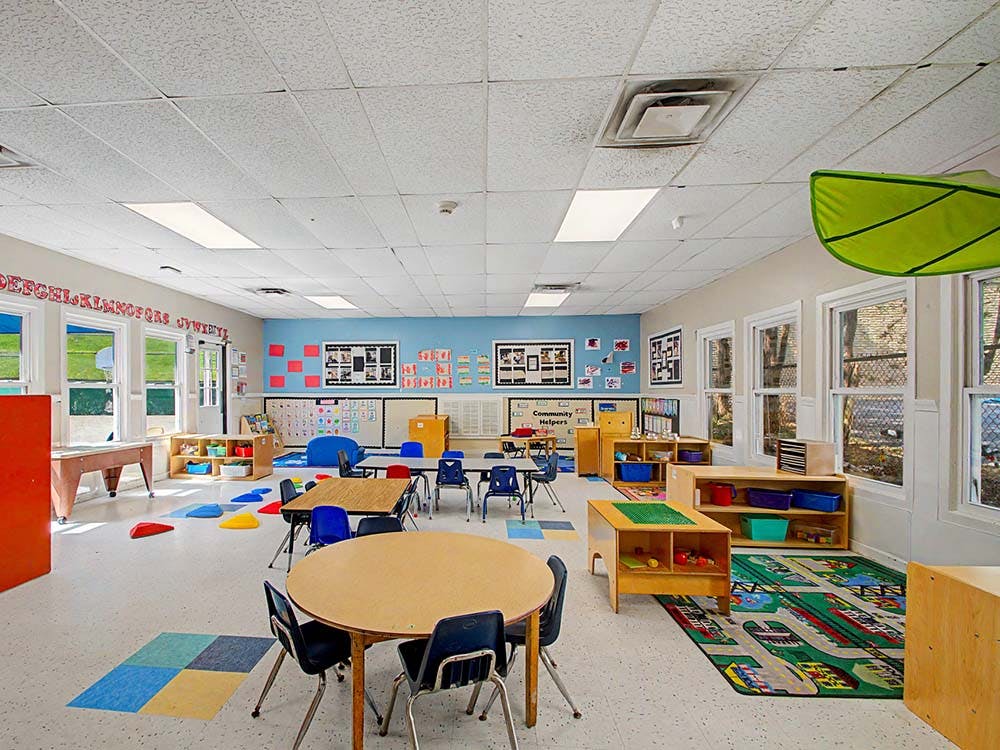 Empty classroom with small tables and chairs, shelves with toys, and alphabet on the wall