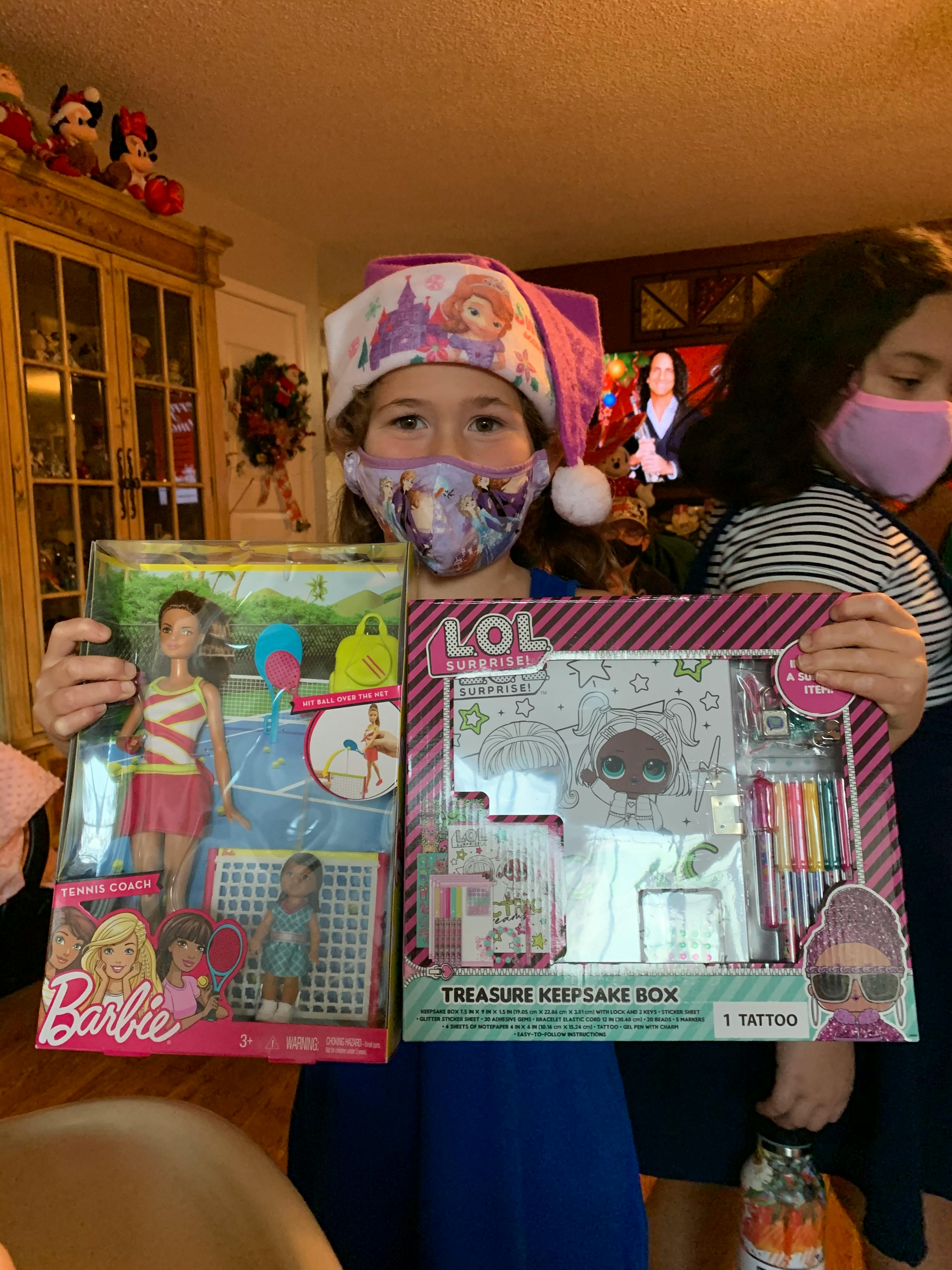 Child in a mask and hat holding a Barbie doll box and an LOL Surprise box