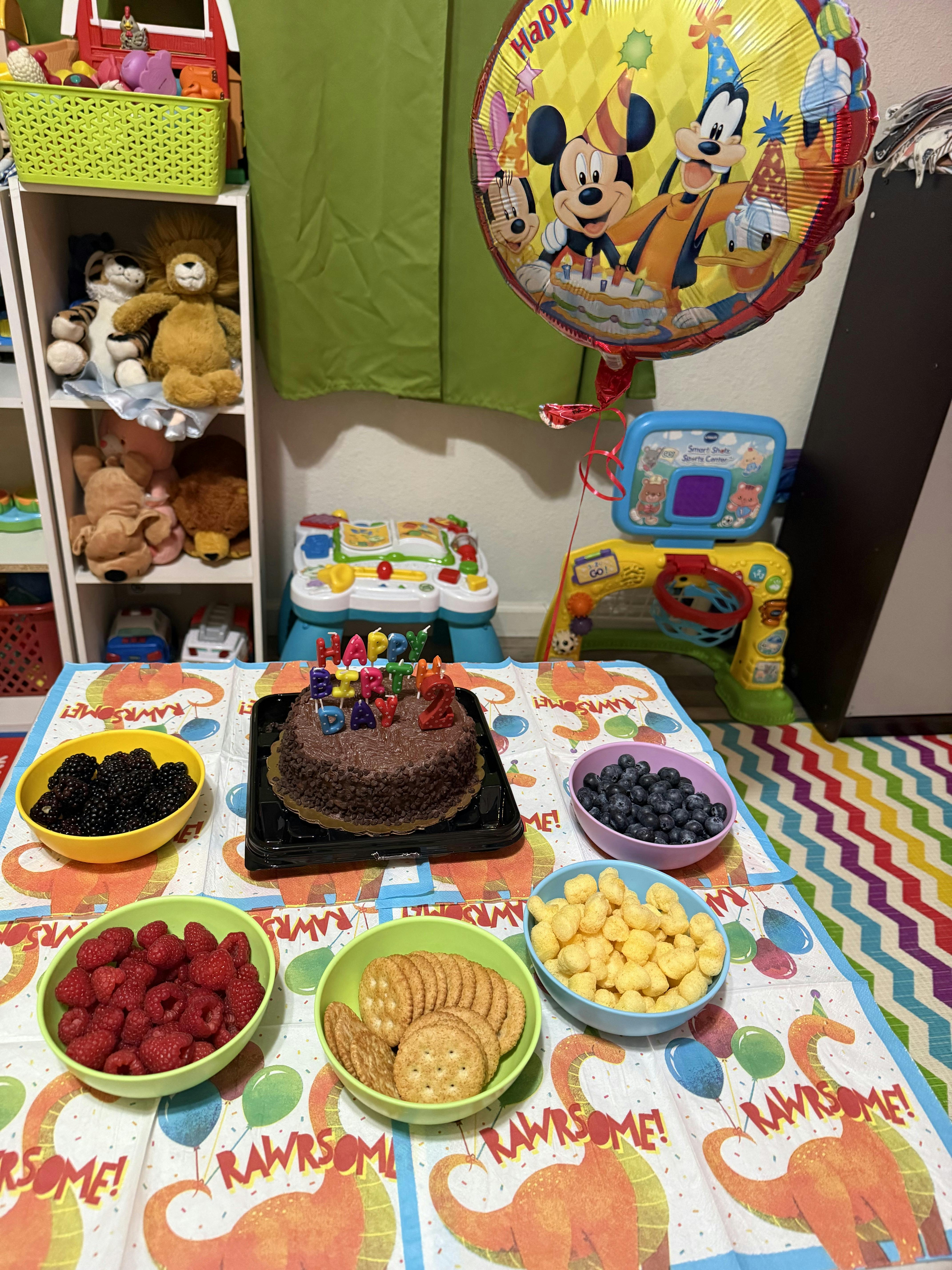 Birthday cake with "Happy Birthday 2" candles, berries, and snacks on a dinosaur tablecloth