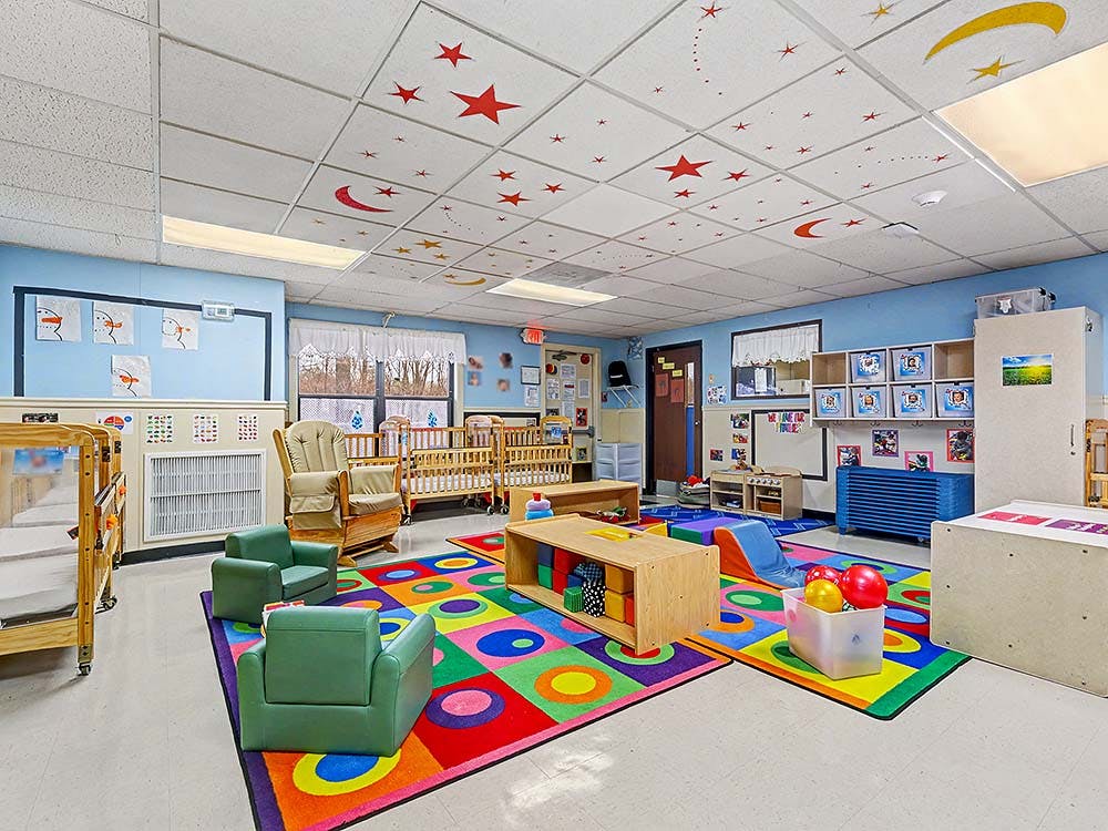 A colorful childcare room with cribs, play mats, and small chairs