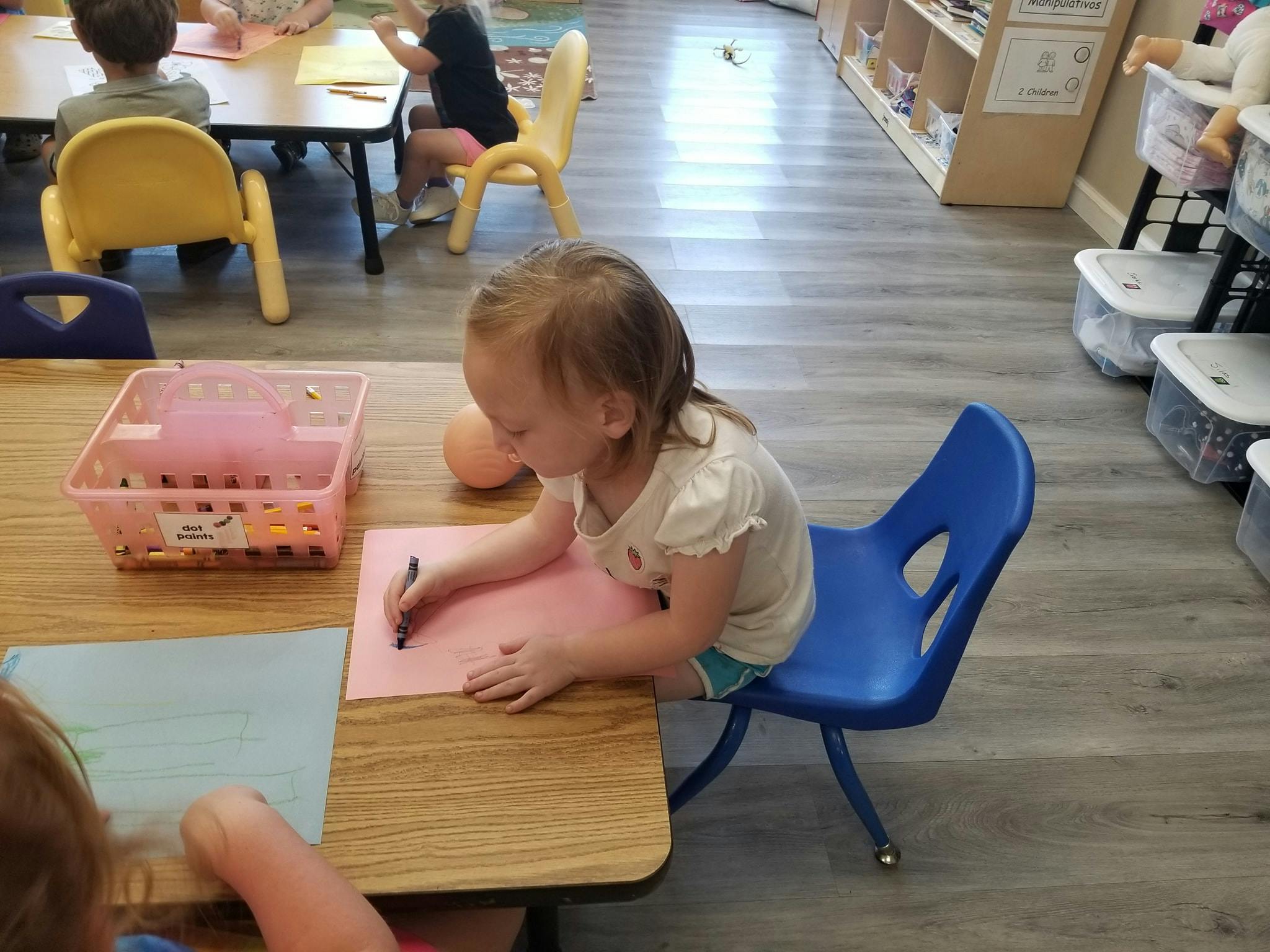 Child sitting at a table drawing on pink paper in a classroom