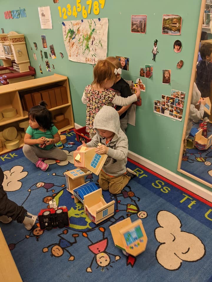 Three children playing in a classroom, one on the floor with blocks, two at a wall