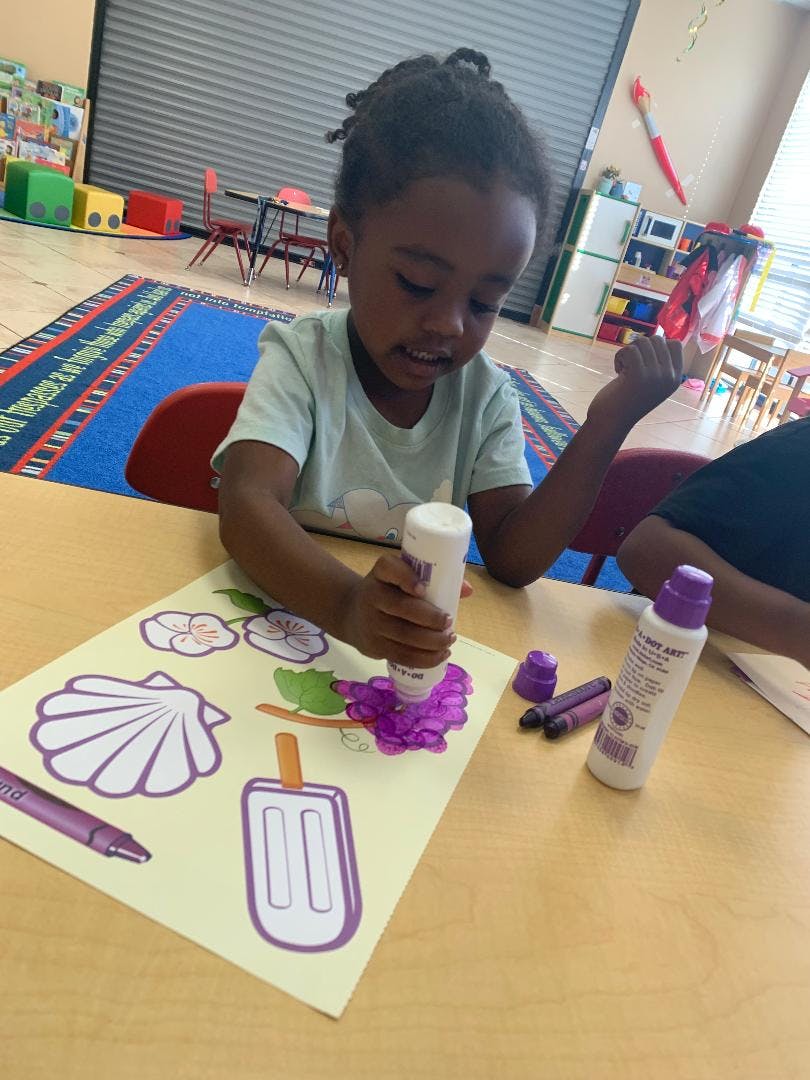 Child applying purple glue to a paper craft in a classroom