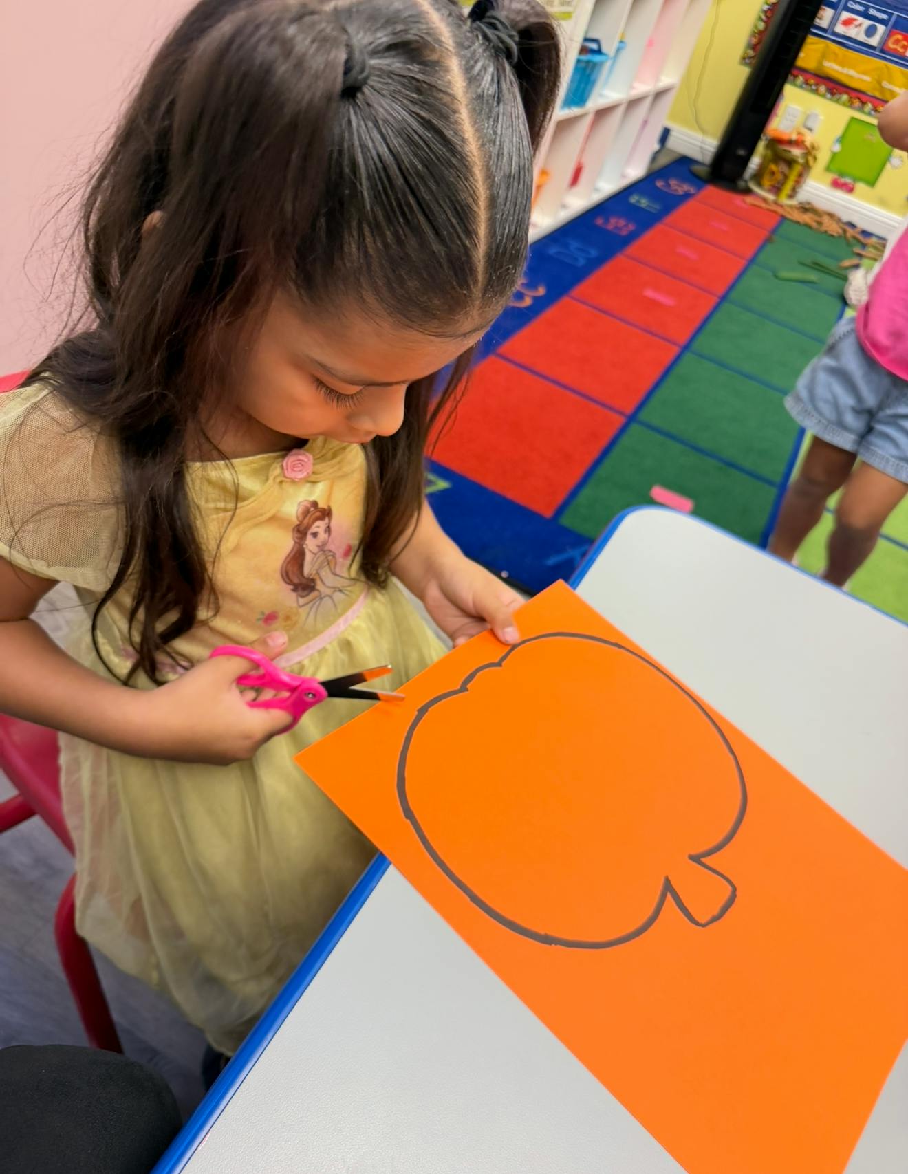Child cutting out a pumpkin shape from orange paper in a classroom