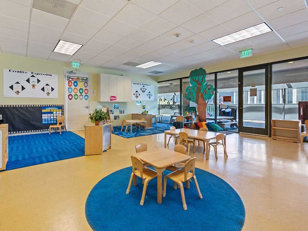 Empty classroom with small tables and chairs, blue rugs, and glass doors