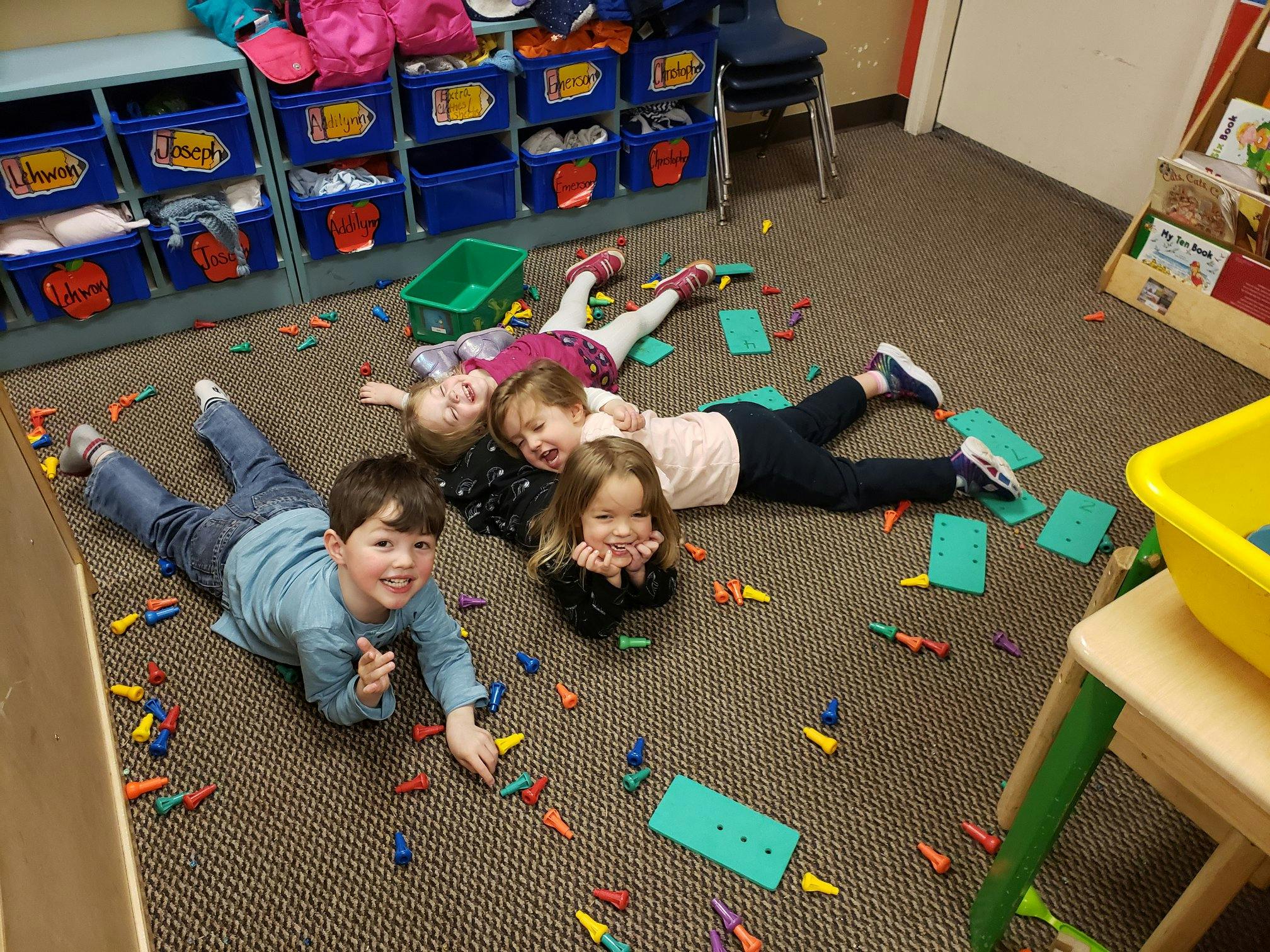 Four children lying on a carpeted floor surrounded by small colorful objects