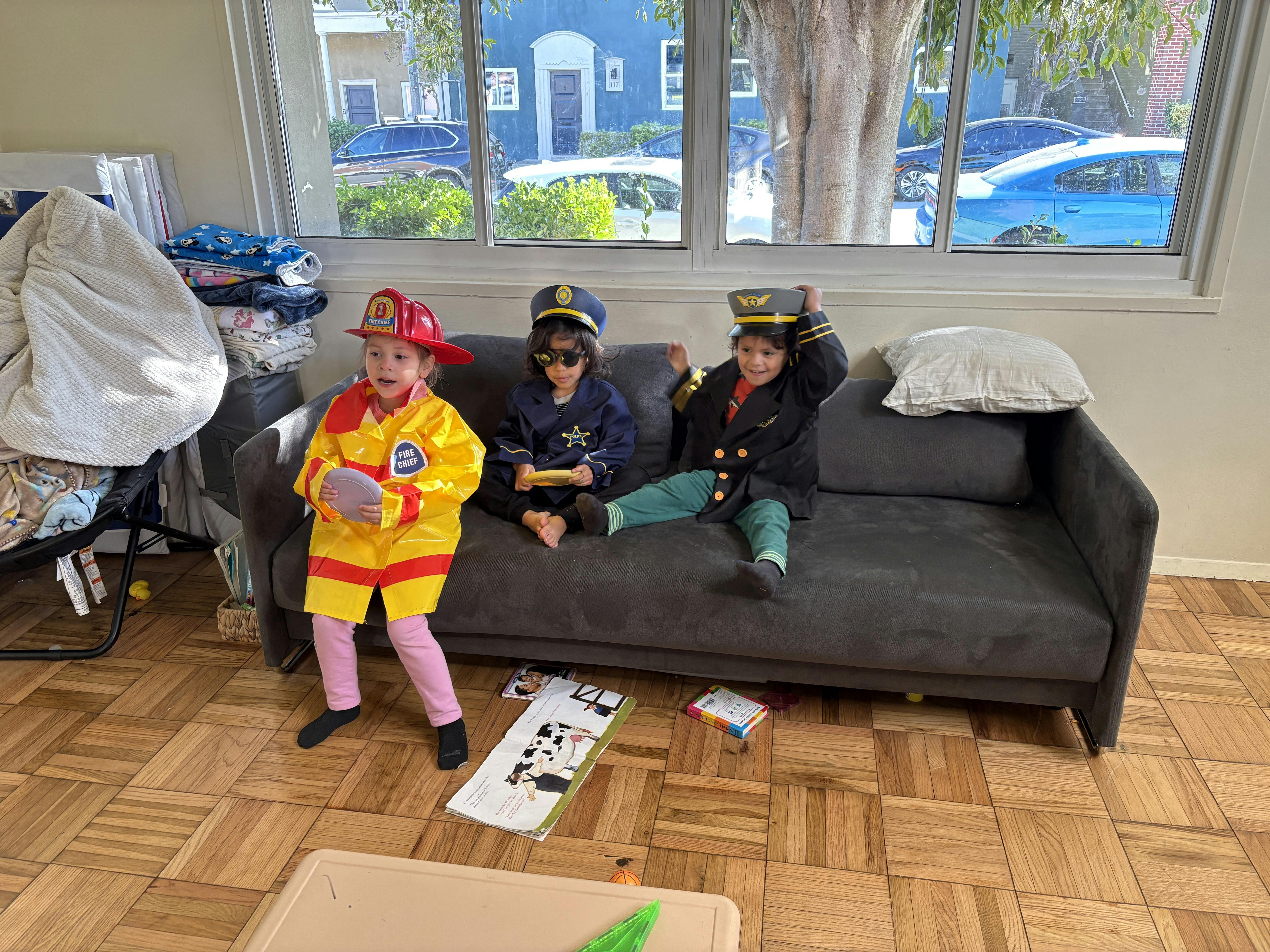 Three children in costumes, one standing in a firefighter costume, two sitting on a couch