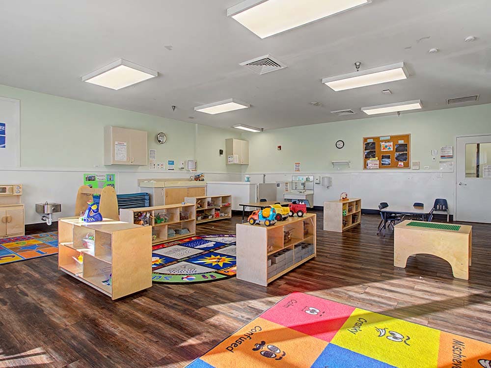 A colorful classroom with play rugs, wooden shelves, and child-sized furniture